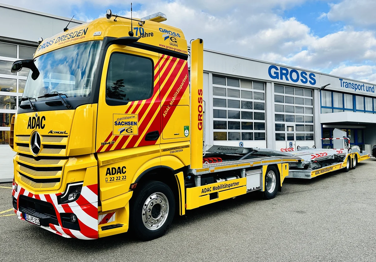 Gelber ADAC Abschleppwagen vor einem Gebäude mit dem Schriftzug GROSS in Blau. Der Lkw ist mit reflektierenden Streifen versehen.