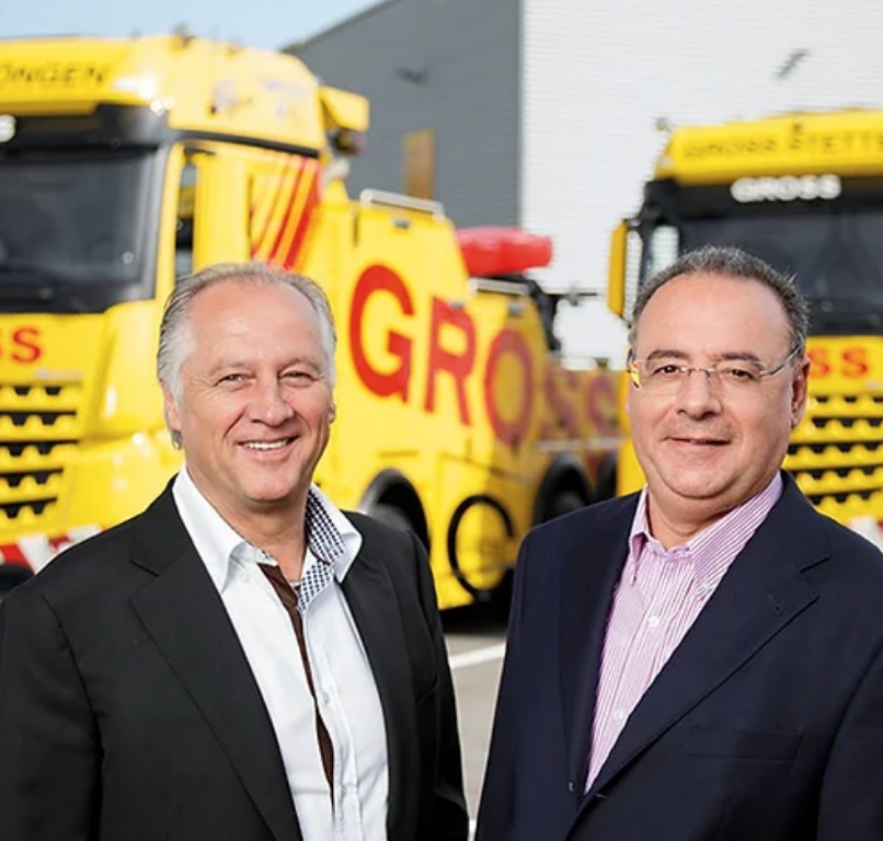 Zwei Männer in Anzügen vor gelben Abschleppwagen. Im Hintergrund sind die Worte GROSS auf den Fahrzeugen zu sehen.