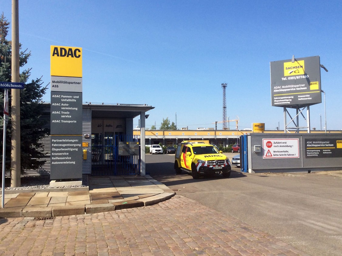 ADAC-Gelände mit gelbem ADAC-Fahrzeug vor Einfahrt, umgeben von Schildern zu Dienstleistungen und Mobilitätspartnern.