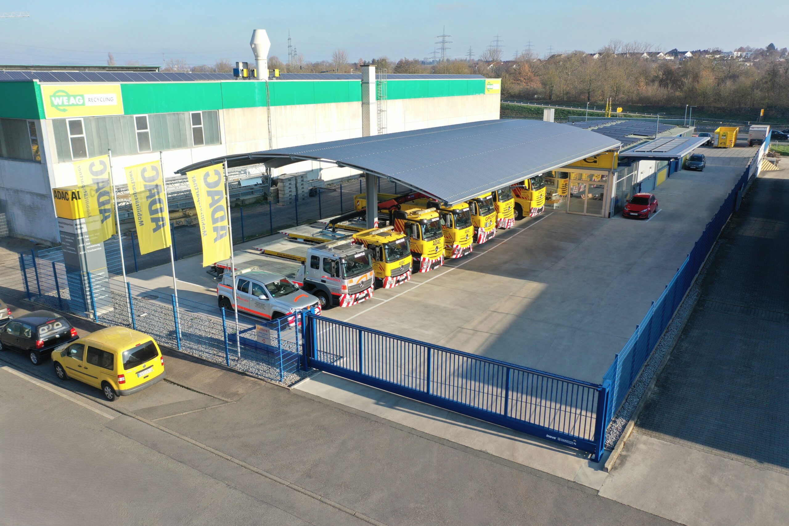 Parkende gelbe Abschleppwagen unter Überdachung bei ADAC-Gebäude, mit weiteren Autos und blauem Zaun im Vordergrund.