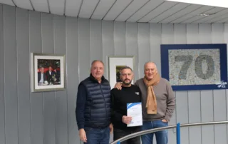 Drei Männer stehen in einem modernen Raum, einer hält eine Urkunde, im Hintergrund sind Bilder und eine 70 sichtbar.
