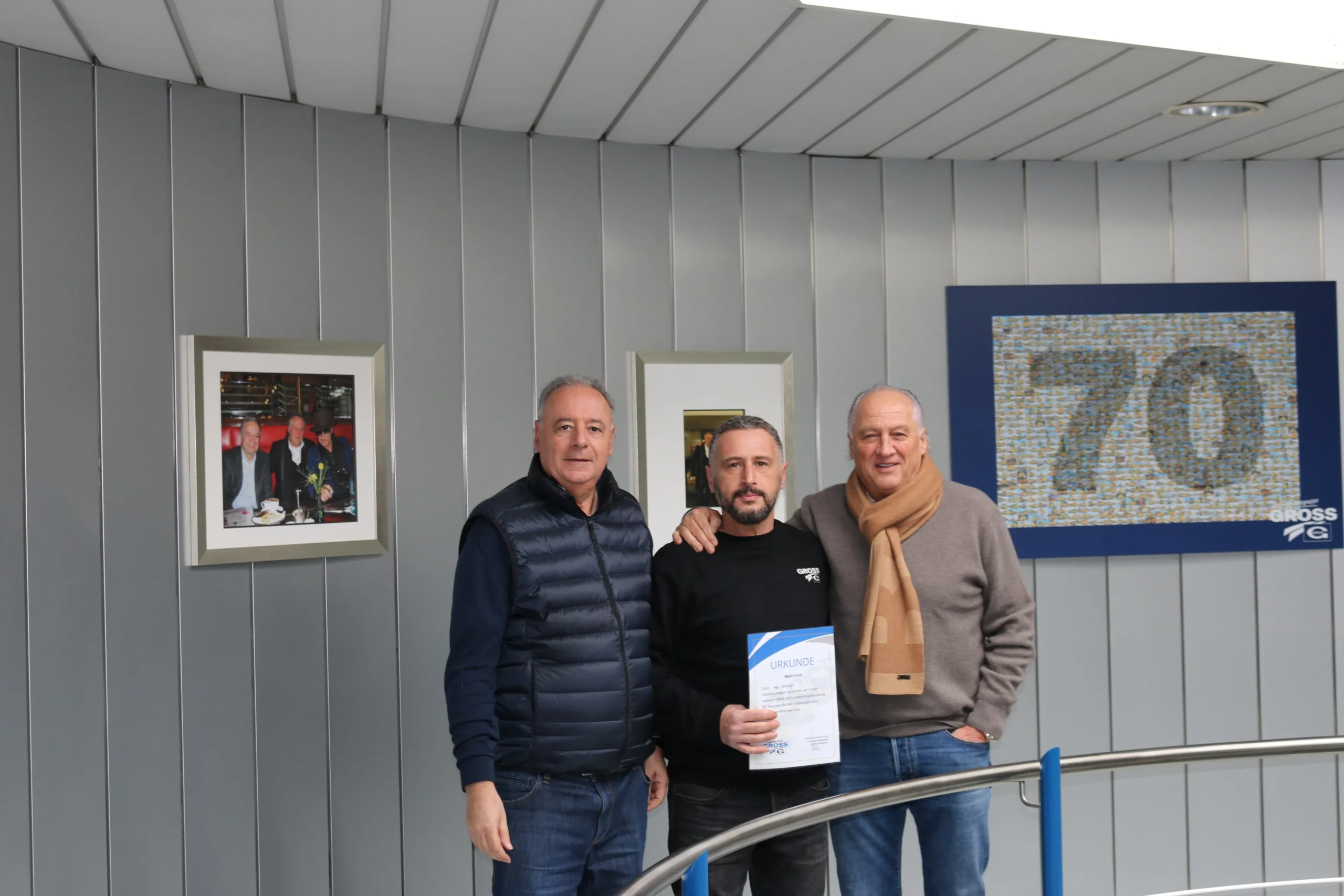 Drei Männer stehen in einem modernen Raum, einer hält eine Urkunde, im Hintergrund sind Bilder und eine 70 sichtbar.