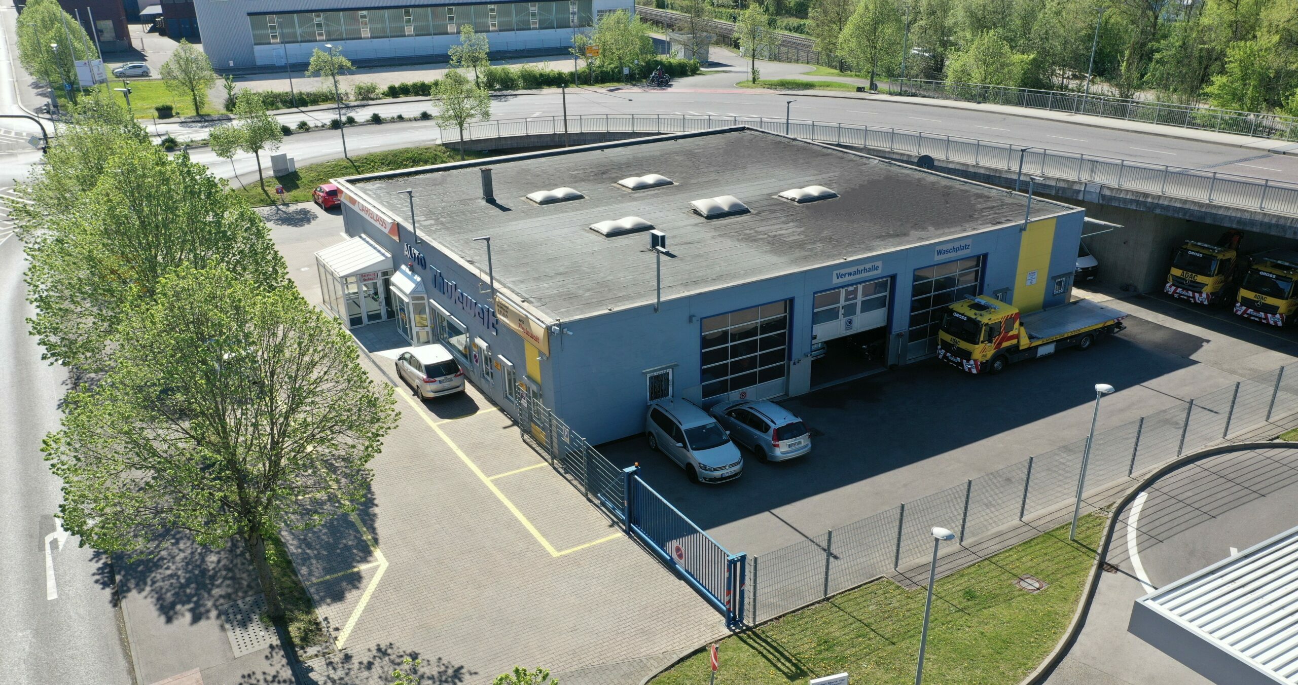 Luftaufnahme eines blauen Industriegebäudes mit Parkplatz, Fahrzeughalle und zwei Abschleppwagen, umgeben von Bäumen und Straßen.