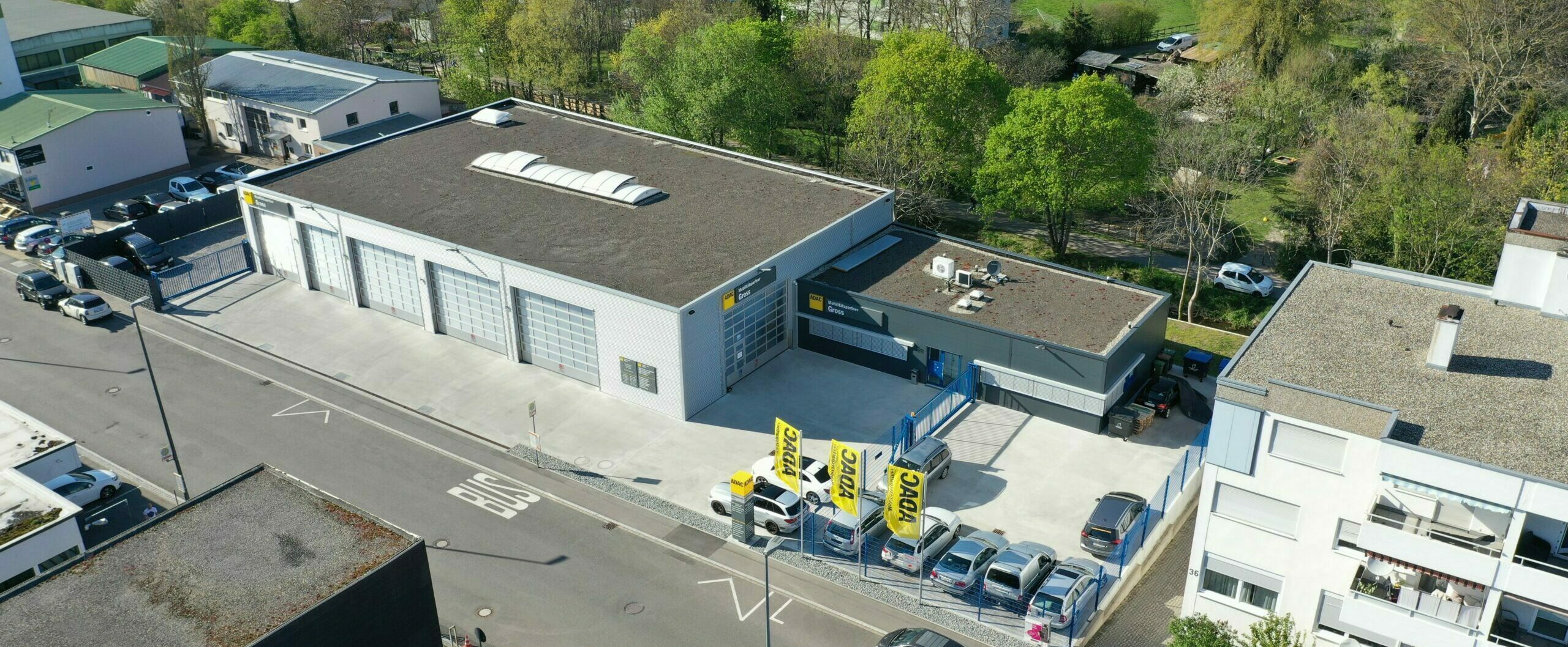 Luftaufnahme einer Werkstatt mit Parkplätzen und ADAC-Flaggen, umgeben von Bäumen und weiteren Gebäuden.
