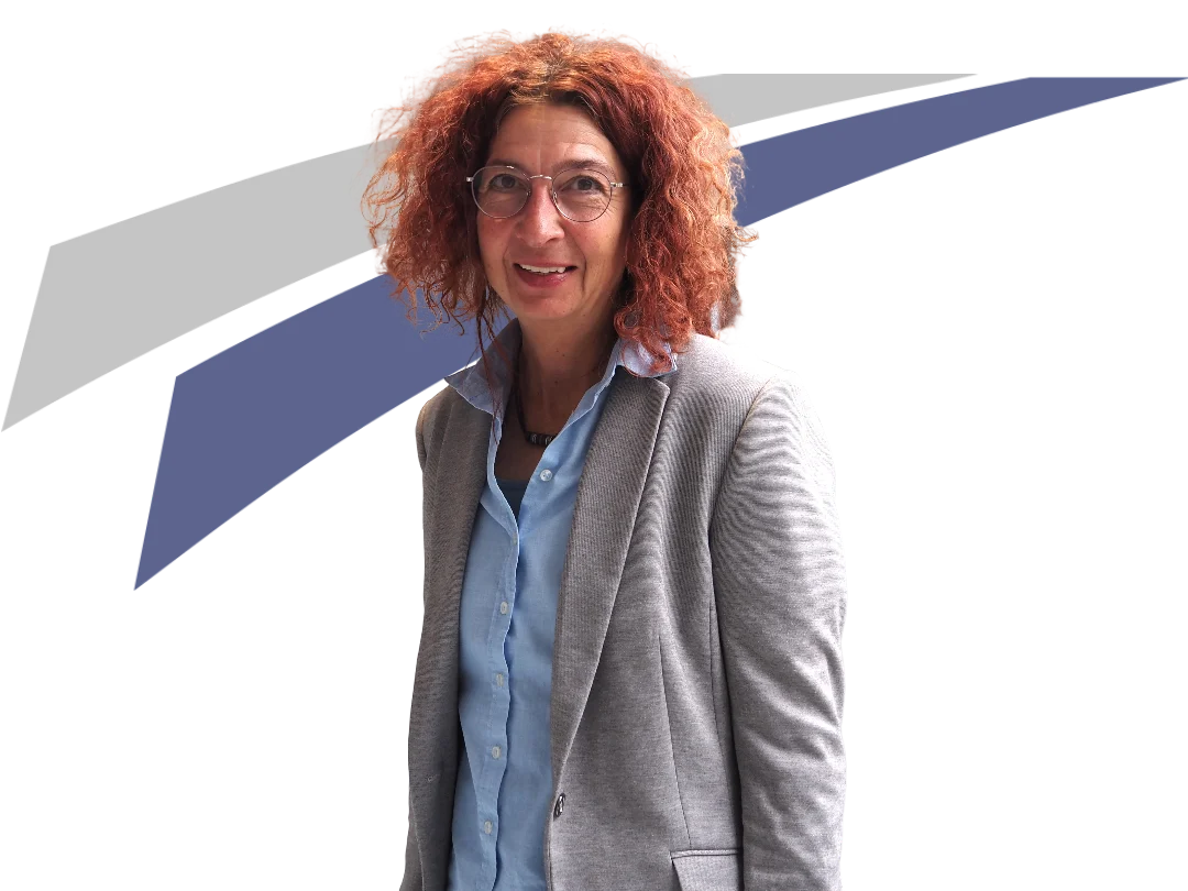 Frau mit lockigem, rotem Haar steht lächelnd vor einem grafischen Hintergrund. Sie trägt eine Brille und einen grauen Blazer.