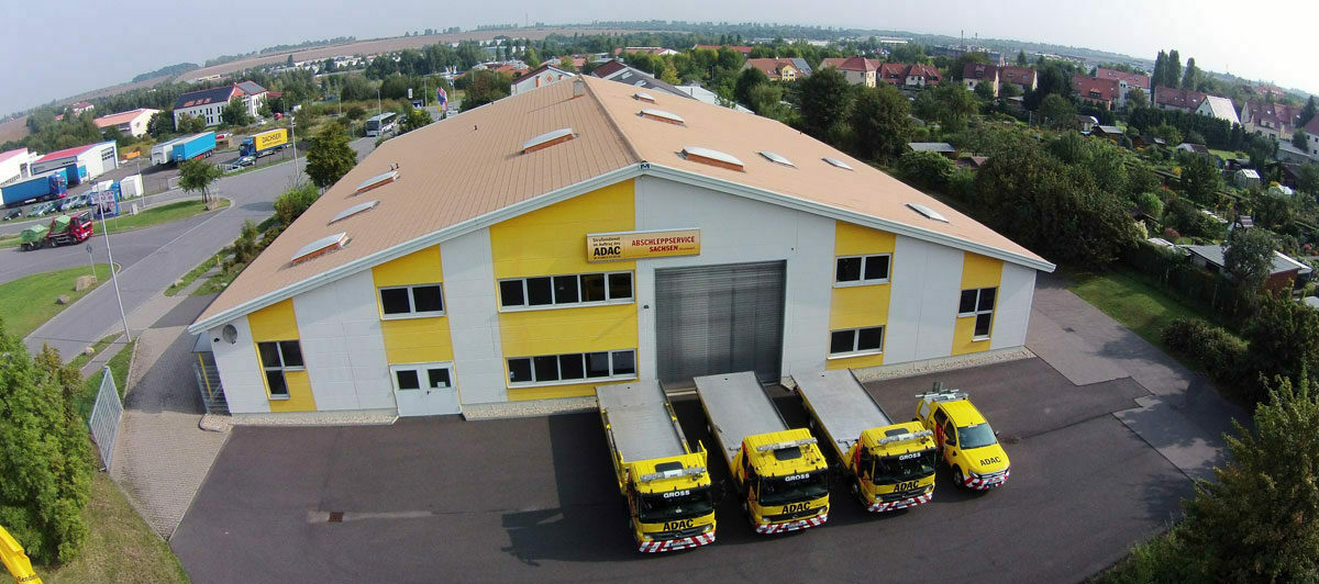 Gelbes Gebäude mit ADAC-Logo und vier Abschleppwagen davor. Umliegende Landschaft und parkende Lastwagen.