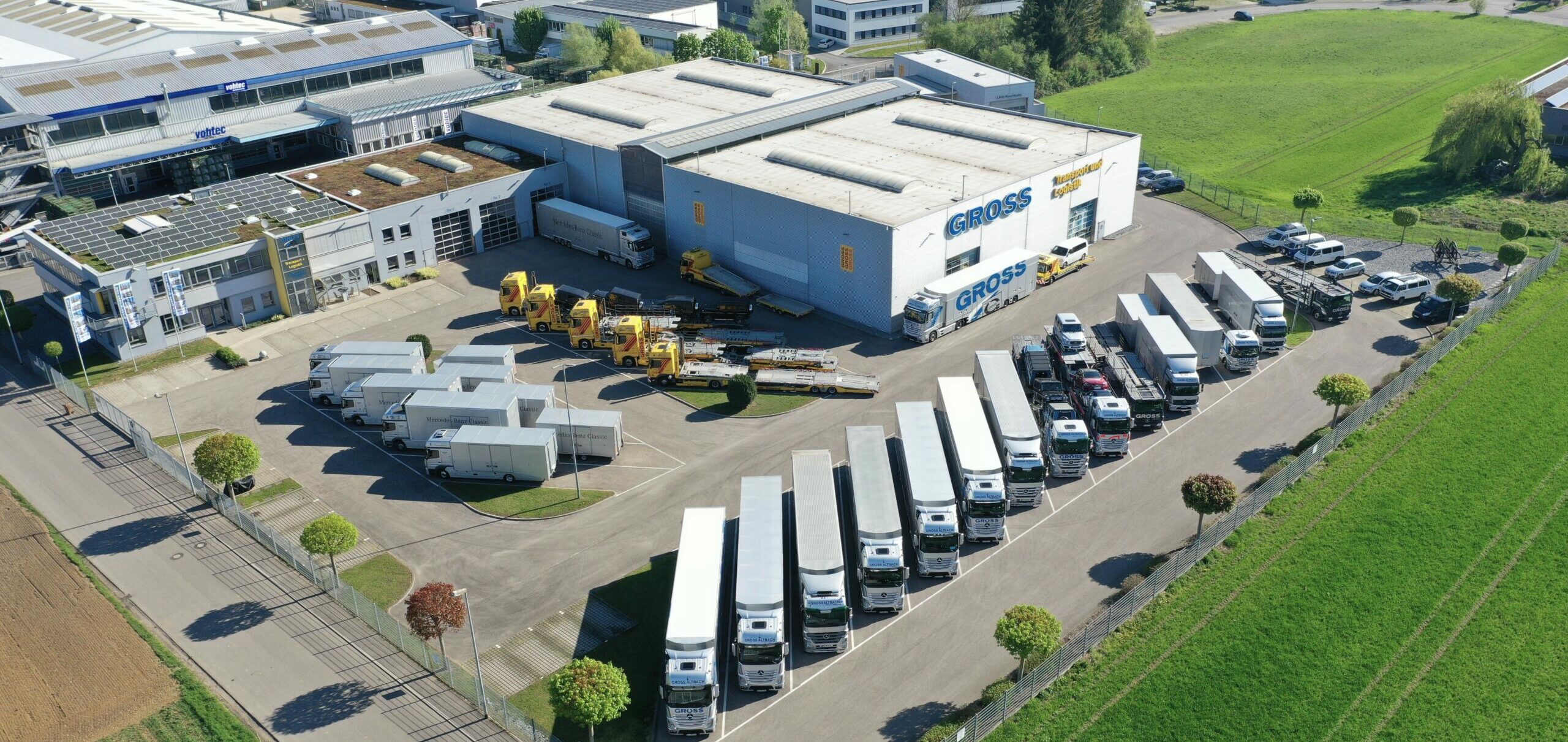 Luftaufnahme eines Logistikzentrums mit mehreren LKWs und Anhängern im Hof neben einem großen Firmengebäude und grünen Feldern.