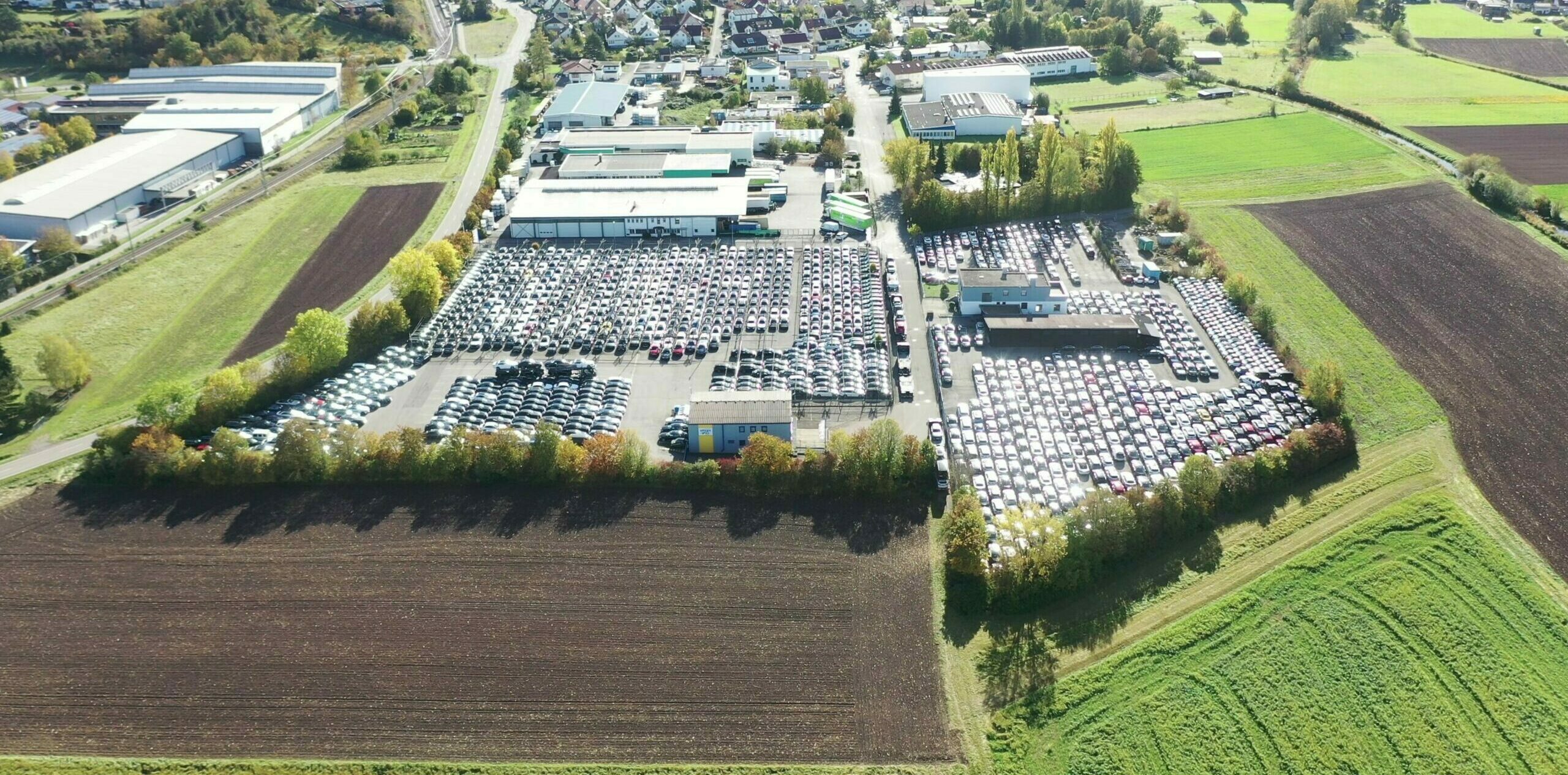 Luftaufnahme eines großen Parkplatzes voller Autos, umgeben von Feldern und Gebäuden in ländlicher Gegend.