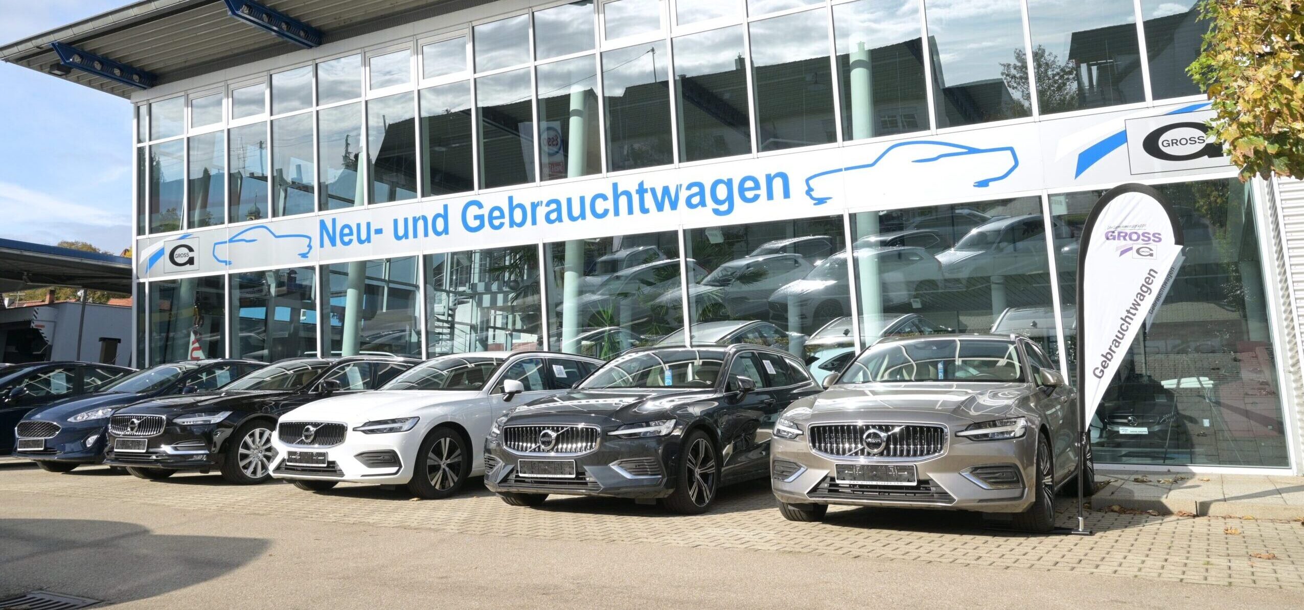 Autohändler mit gebogenen Fahrzeugen vor Glasfassade. Banner: Neu- und Gebrauchtwagen.