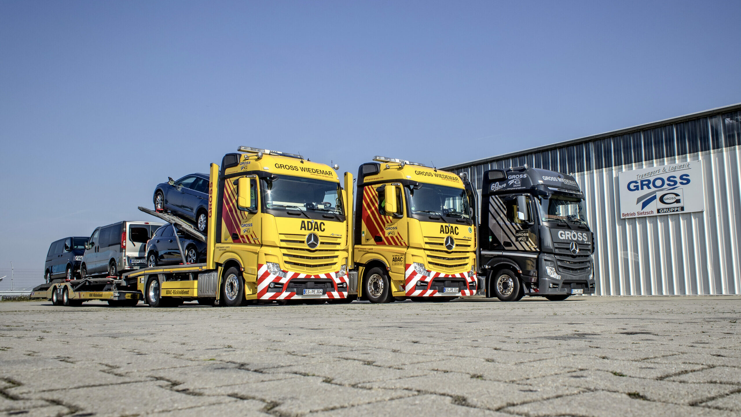 Gelbe ADAC-Abschleppwagen mit Autos beladen, stehen auf einem Parkplatz neben einem schwarzen Lkw vor einer Lagerhalle.