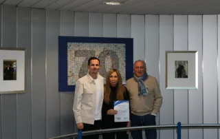 Drei Personen stehen an einem Geländer und halten gemeinsam eine Urkunde vor einem bunten Kunstwerk an der Wand.