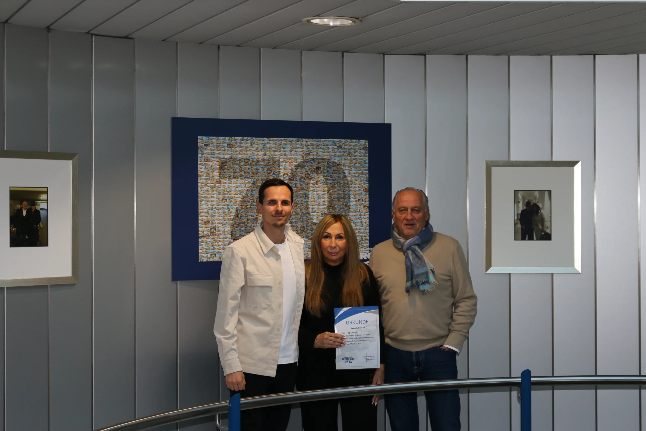 Drei Personen stehen an einem Geländer und halten gemeinsam eine Urkunde vor einem bunten Kunstwerk an der Wand.