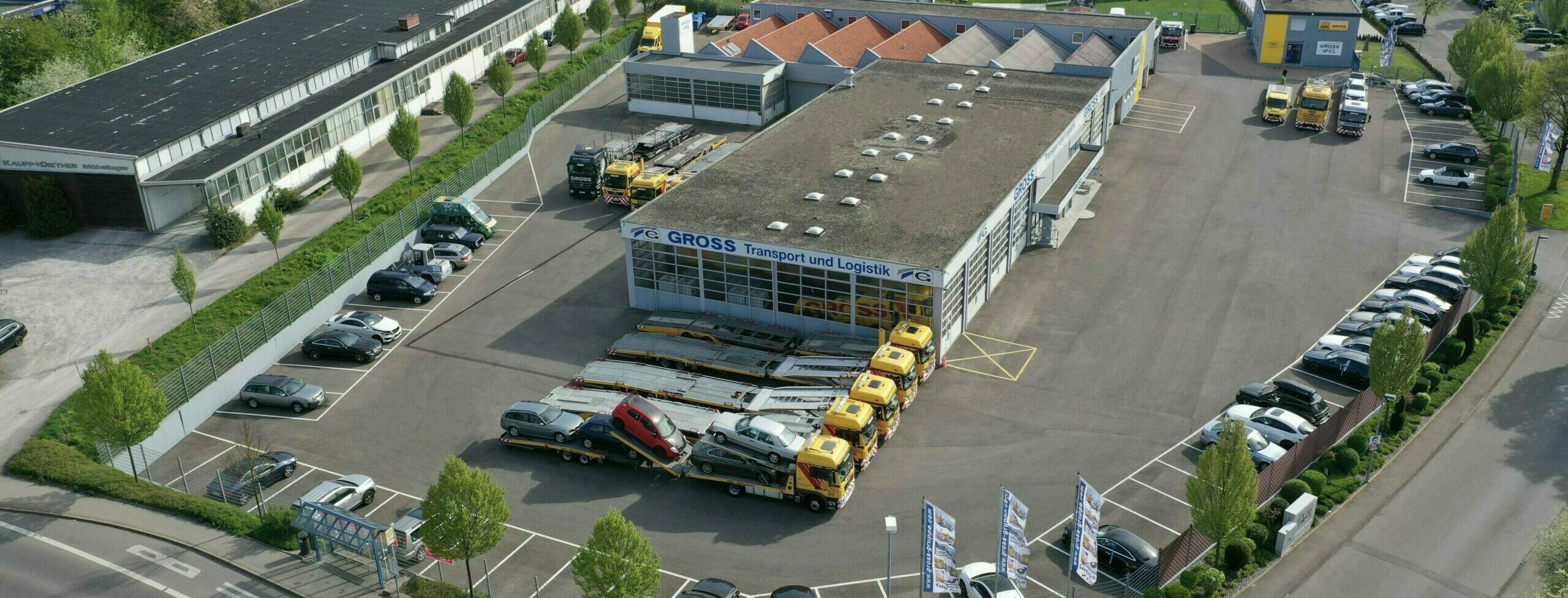 Luftaufnahme eines Logistikzentrums mit parkenden Transportern und Autos auf einem großen Parkplatz.
