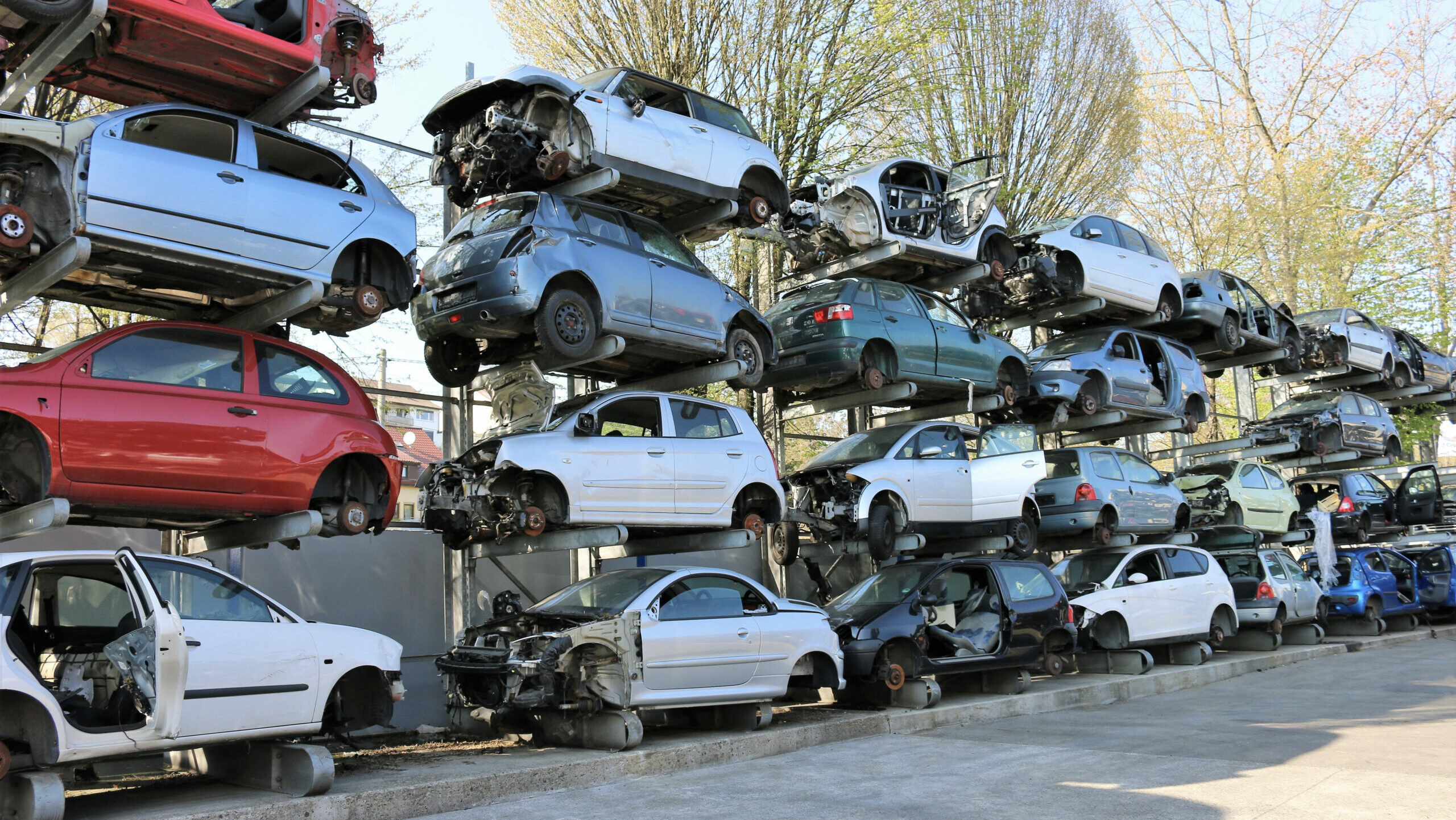 Gestapelte Schrottautos ohne Reifen auf einem Autoverwertungsplatz, umgeben von Bäumen im Hintergrund.