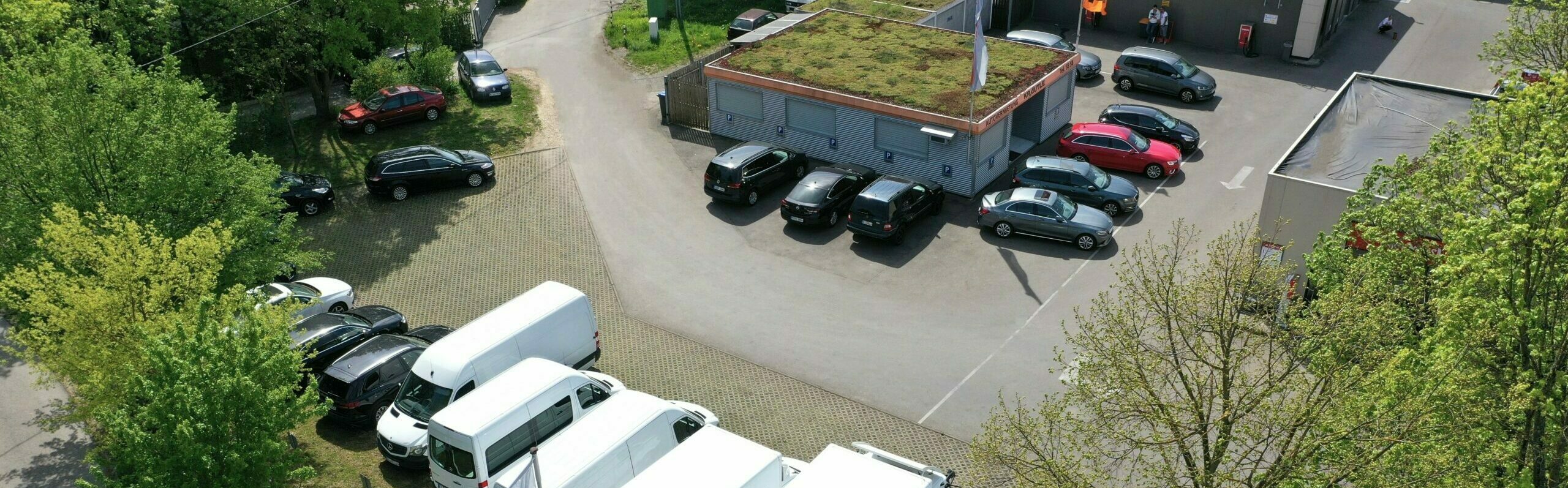 Luftaufnahme eines Parkplatzes mit Autos und weißen Transportern neben einem Gebäude mit begrüntem Dach, umgeben von Bäumen.