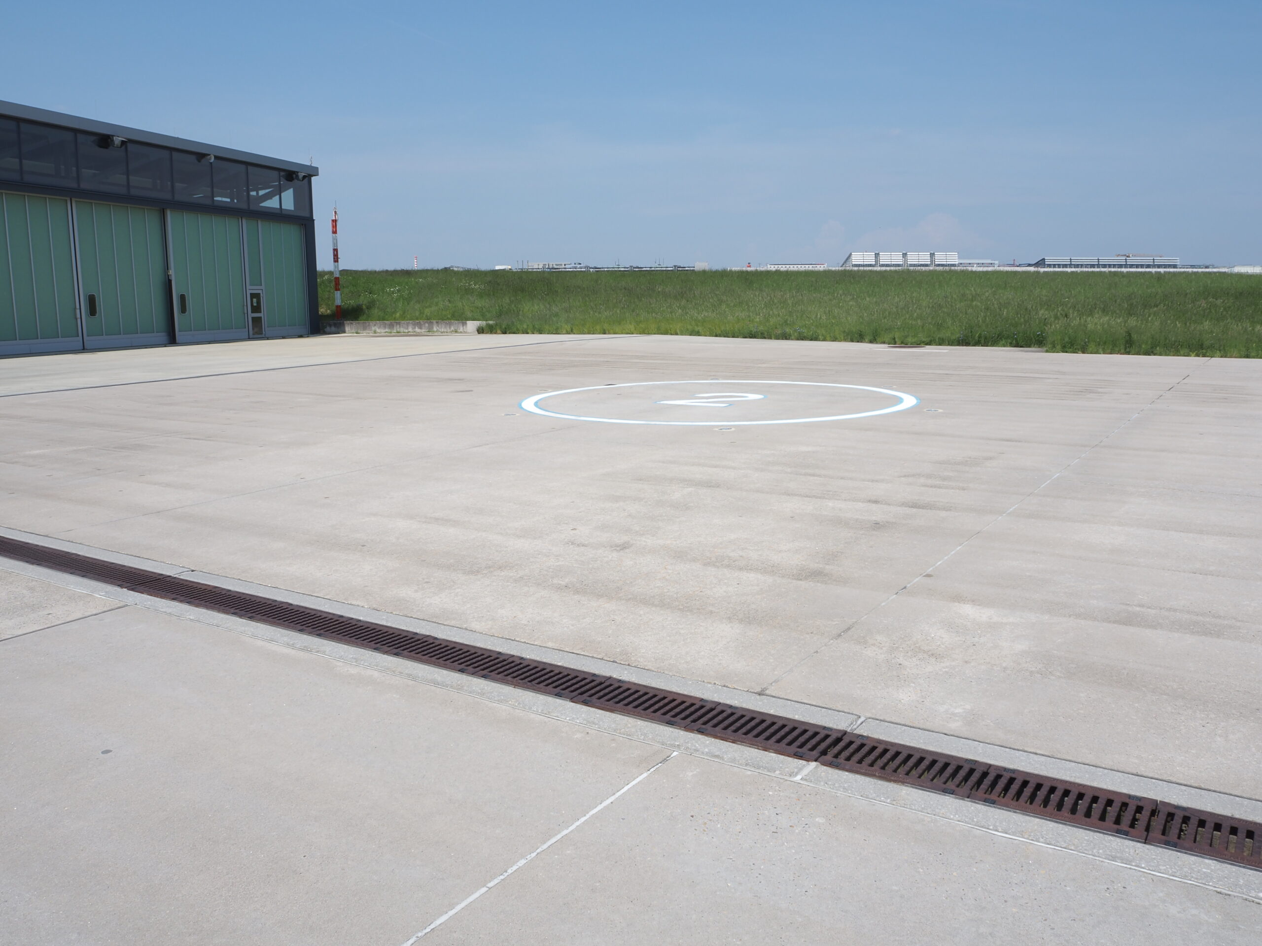 Hubschrauberlandeplatz neben einem modernen Hangar, umgeben von grünem Gras und klarem, blauem Himmel im Hintergrund.