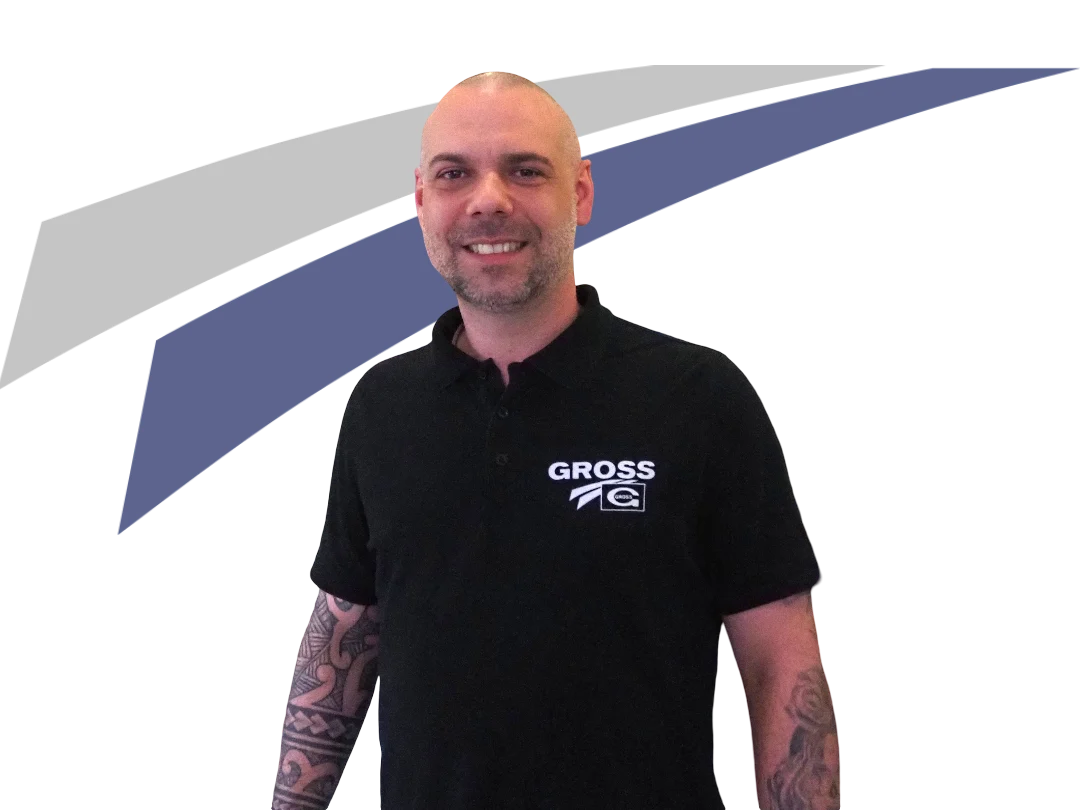 Mann mit tätowierten Armen lächelt in schwarzem Poloshirt mit Logo vor einem grafischen, graublauen Hintergrund.