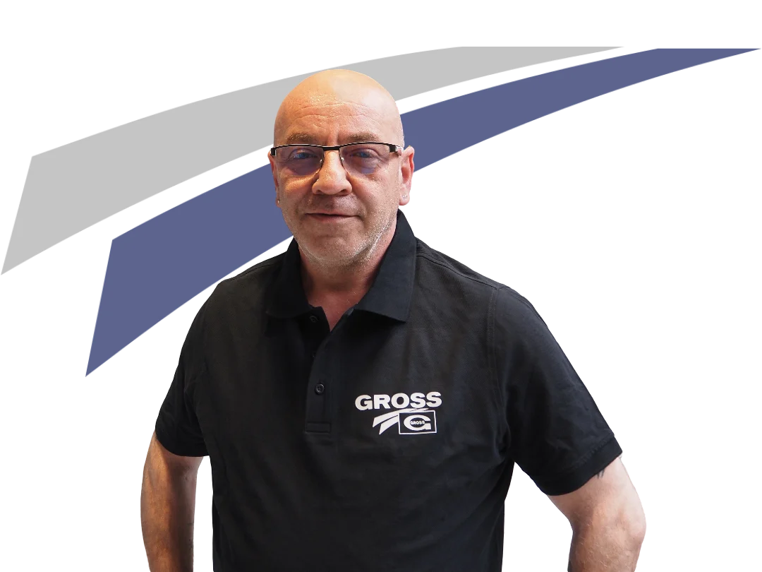 Mann mit Glatze, Brille, schwarzem Hemd mit GROSS-Logo, steht vor einem grauen und blauen Hintergrund.