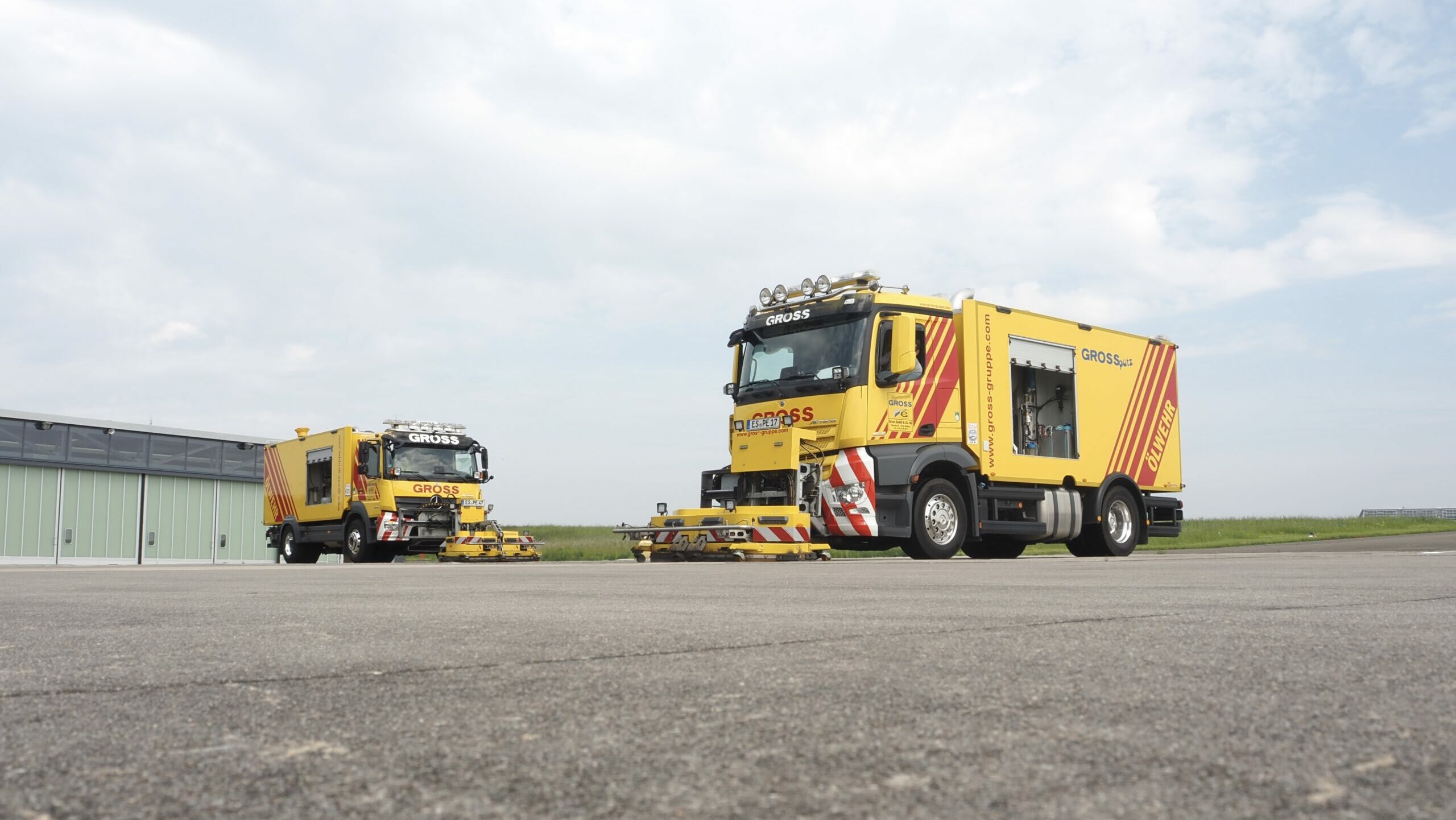 Zwei gelbe Ölwehrfahrzeuge mit roten Streifen auf einer Asphaltfläche, bereit für den Einsatz vor einem Gebäude.