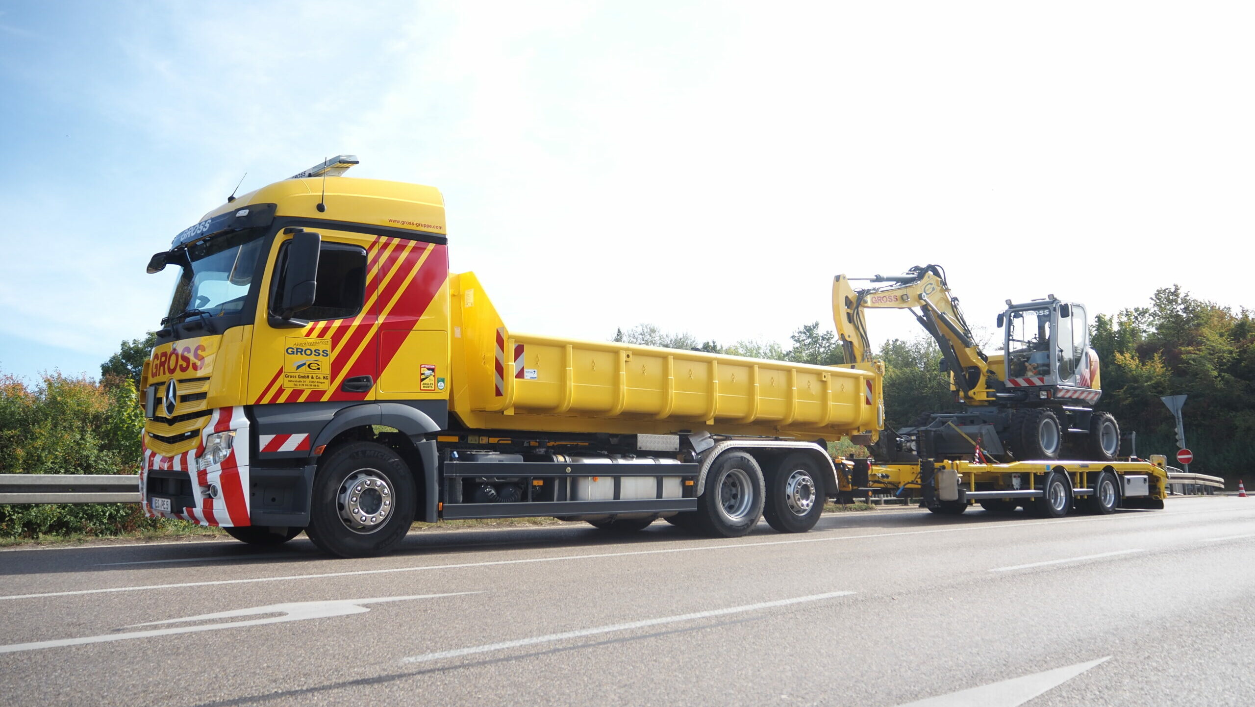 Gelber Lastwagen mit tiefer Ladefläche transportiert einen kleinen Bagger auf einer Landstraße bei Sonnenschein.