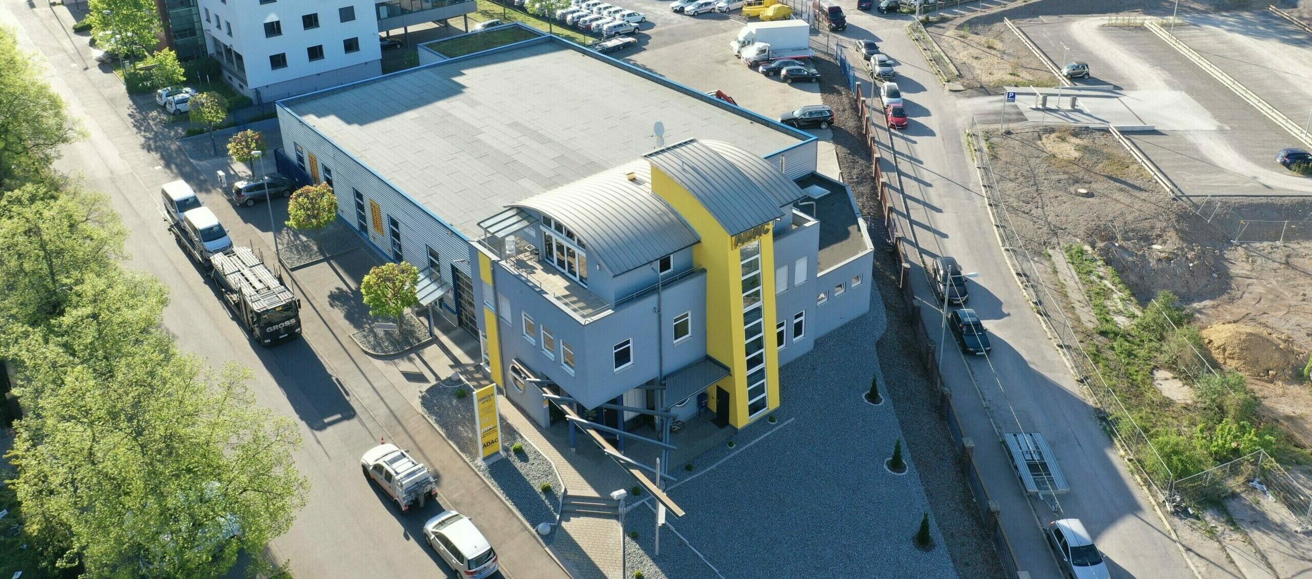 Luftaufnahme eines modernen grauen Gebäudes mit gelben Akzenten, umgeben von Straßen und Autos, bei sonnigem Wetter.