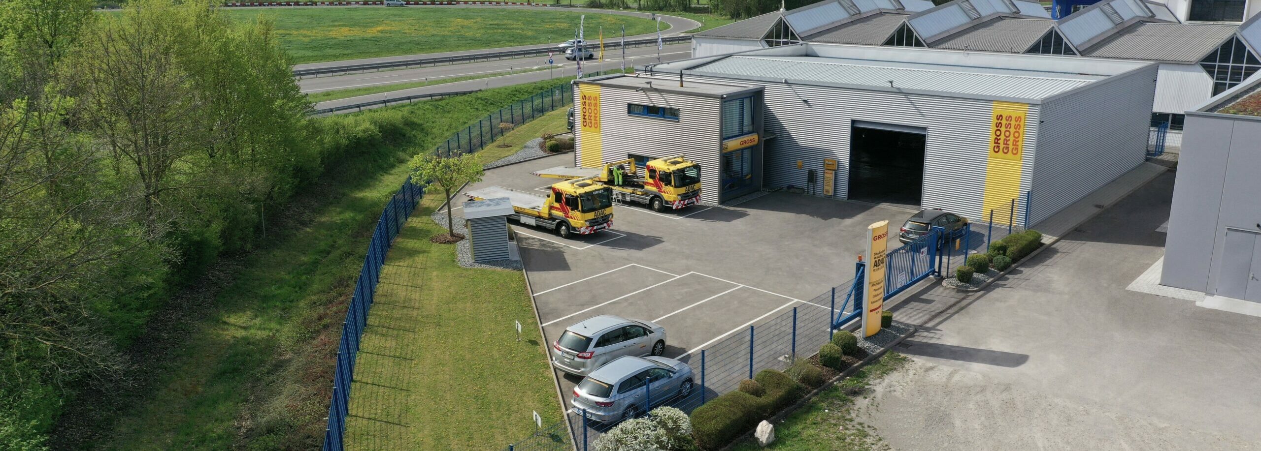 Gewerbegebäude mit drei Abschleppwagen auf Parkplatz, umgeben von Bäumen und Zaun, nahe einer Straße.