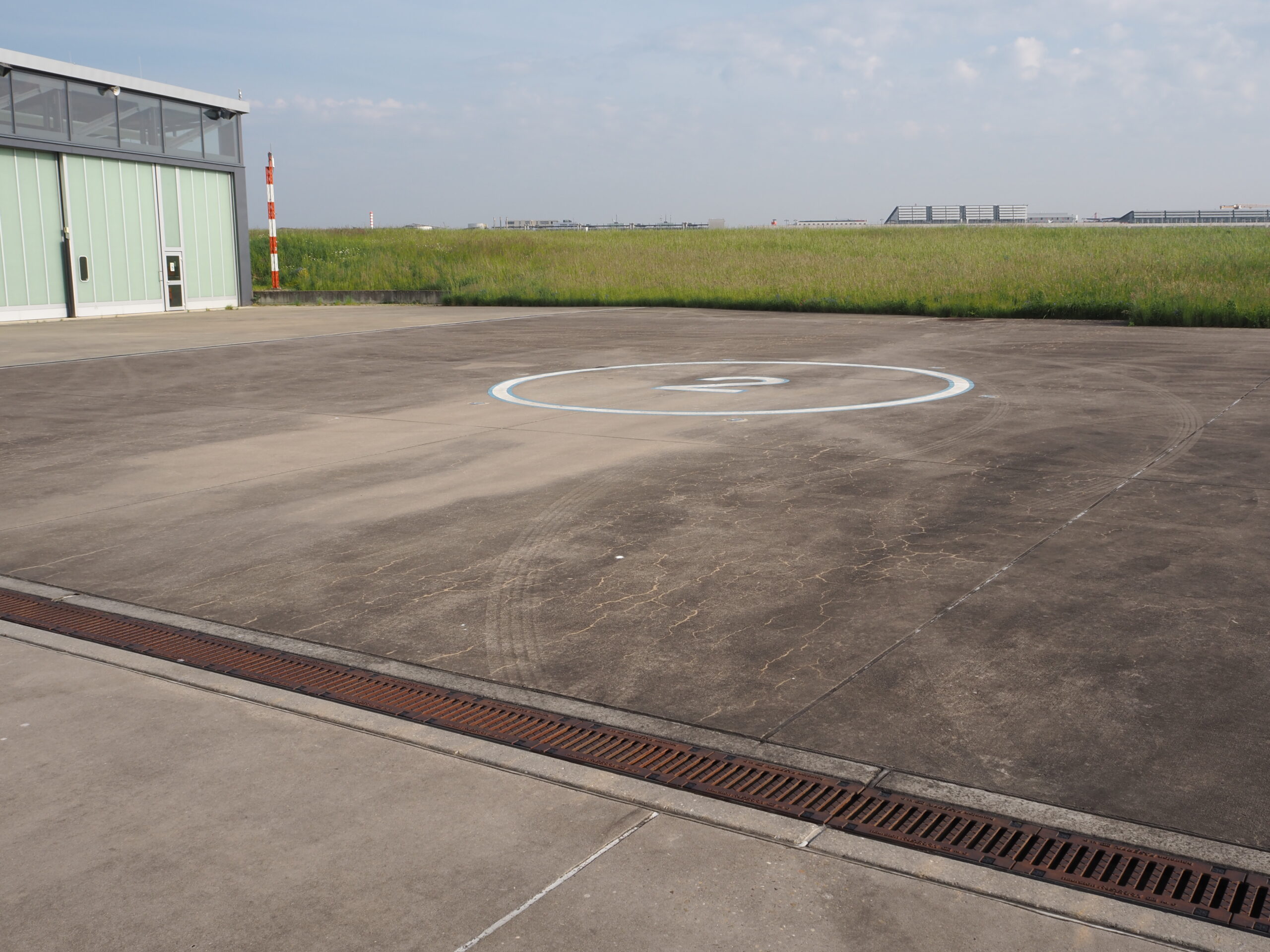Helipad auf leerem Gelände neben Gebäude, umgeben von Gras und Himmel im Hintergrund.