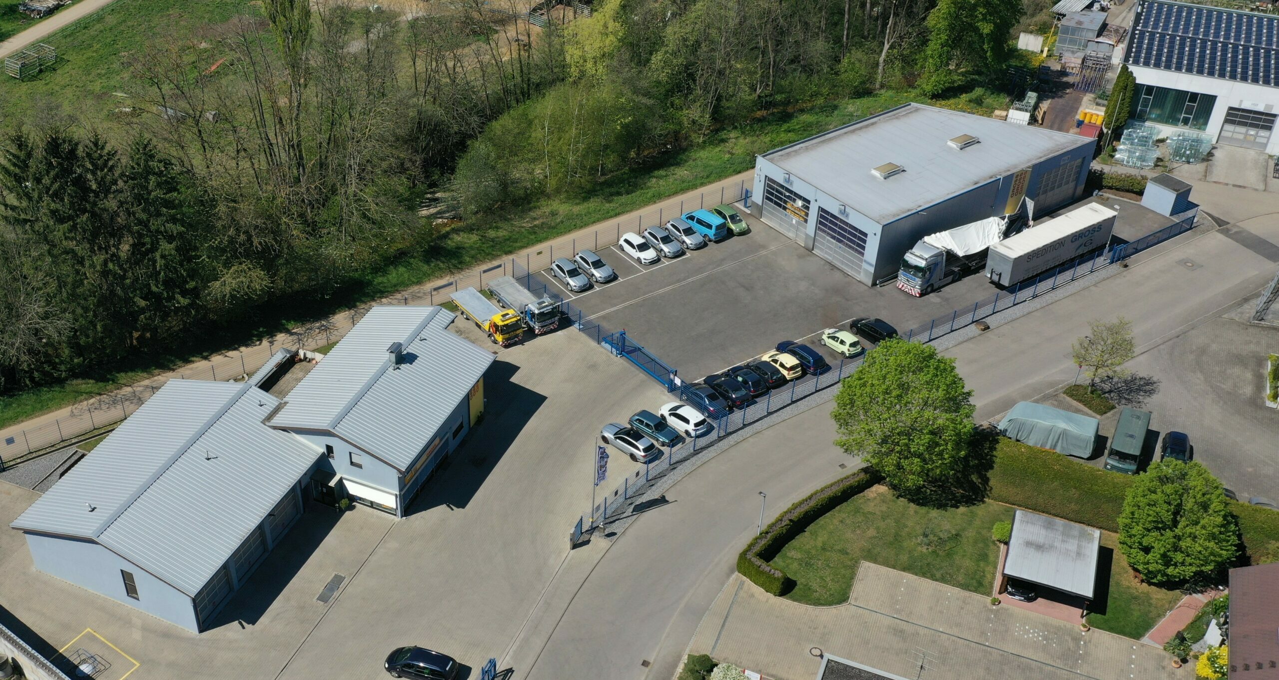 Luftaufnahme eines Industriegebäudes mit Parkplatz, LKW und Autos, umgeben von Bäumen und grüner Fläche.