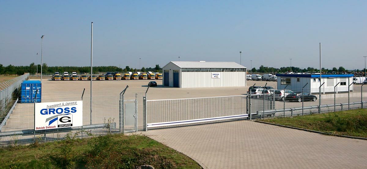 Logistikzentrum mit parkenden Lastwagen und Gebäuden hinter einem Zaun, im Fokus stehen Trucks und Bürocontainer.
