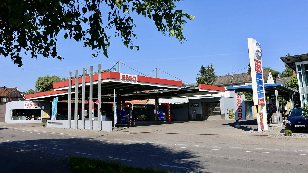 Tankstelle mit mehreren Zapfsäulen, einem roten Dach und einem Shop. Im Vordergrund Bäume und Straße bei Tageslicht.