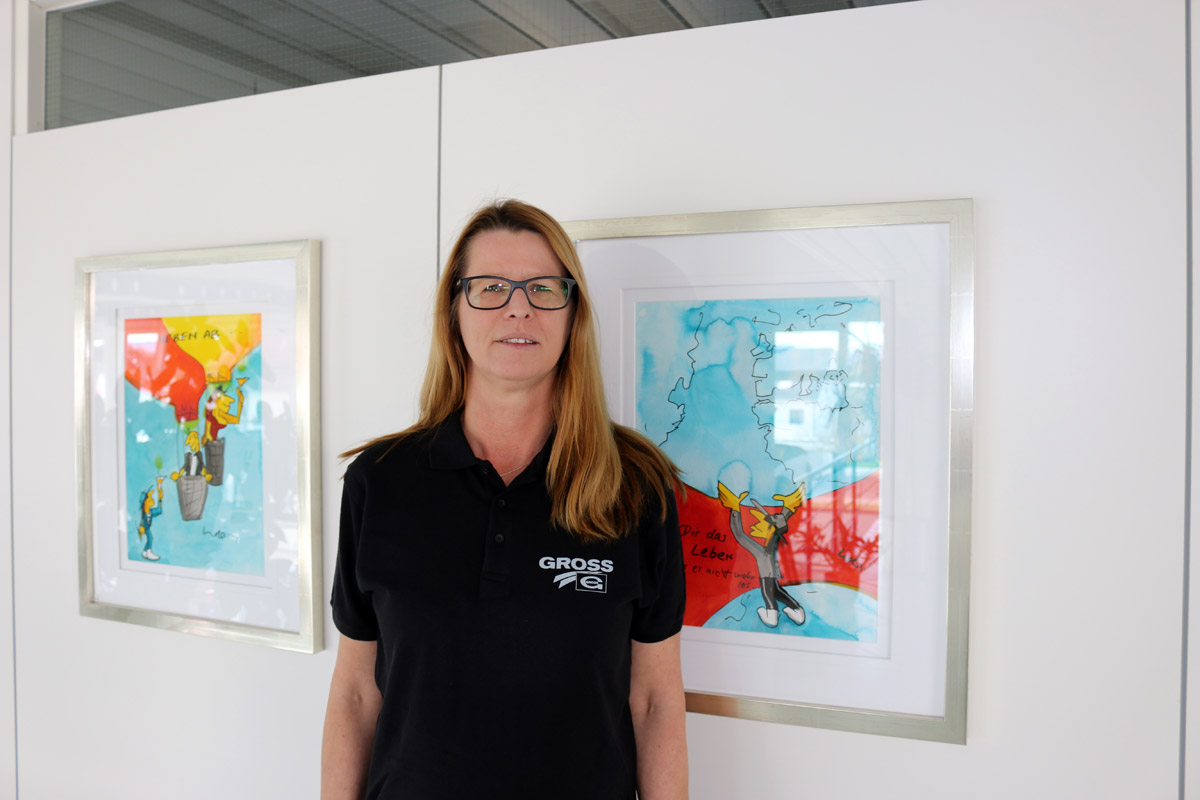 weilheim-natterer-xl Frau mit Brille in schwarzem Polohemd steht vor zwei bunten, gerahmten Cartoon-Illustrationen an einer weißen Wand.