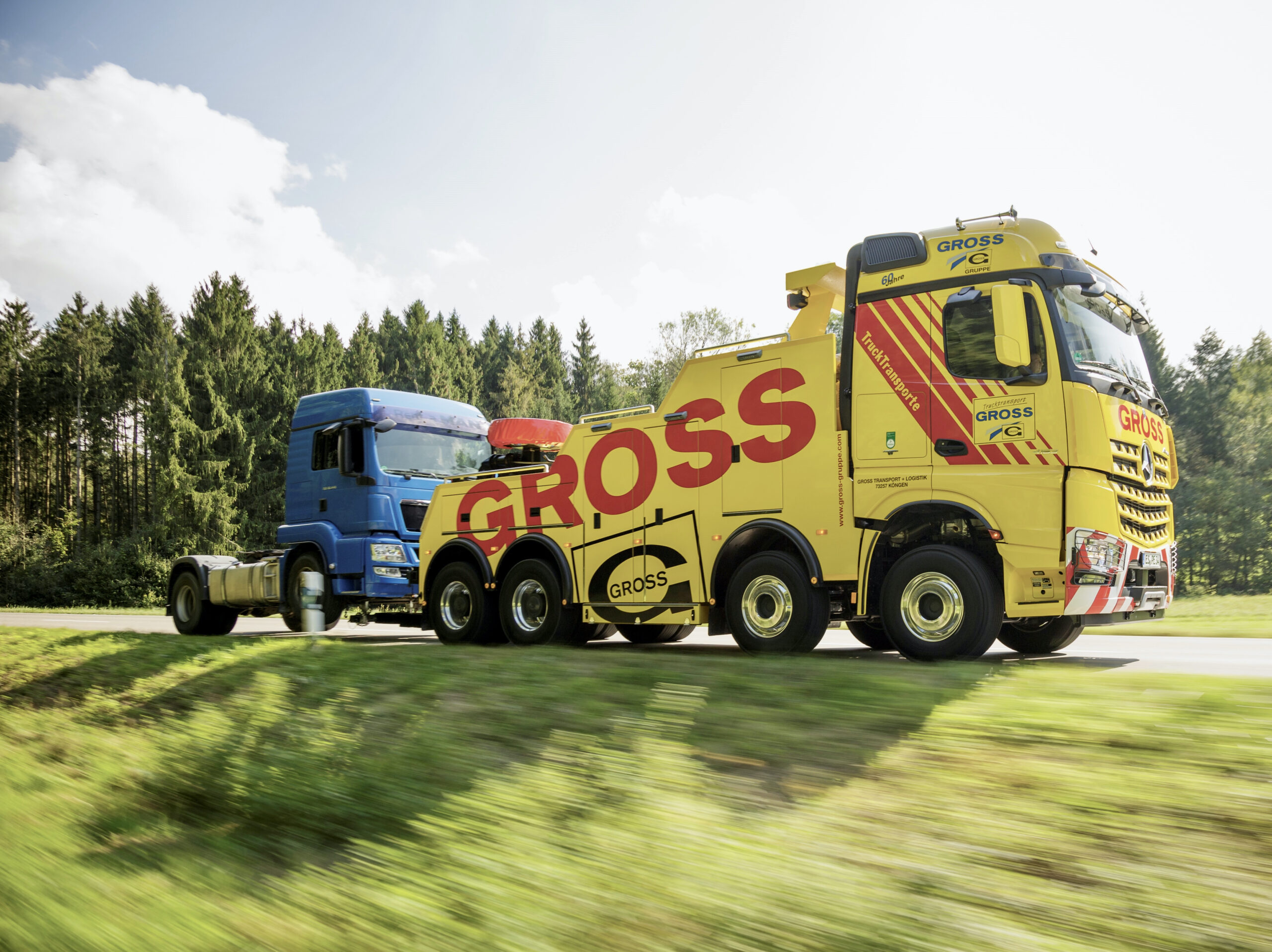 Gelber Abschleppwagen mit Gross-Aufschrift zieht blauen Lkw vor bewaldetem Hintergrund über eine Landstraße.