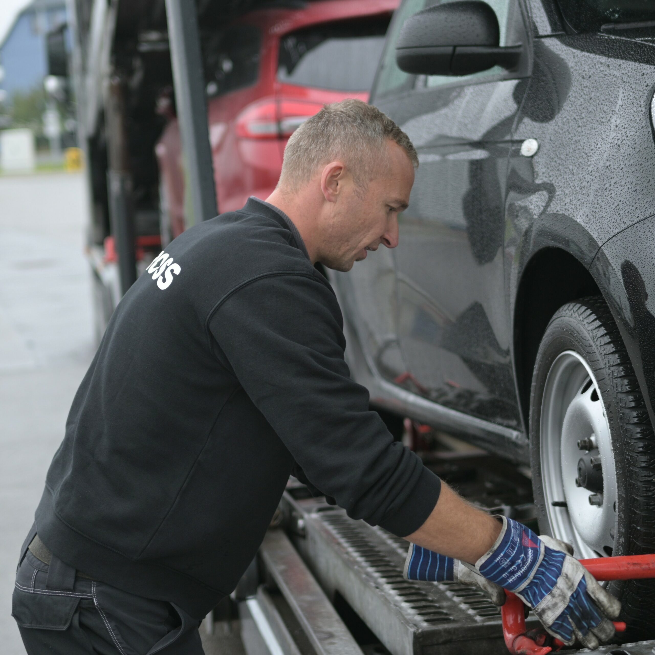 Mann befestigt schwarzes Auto auf Autotransporter mit nassen Reifen, trägt schwarze Jacke und blaue Handschuhe.
