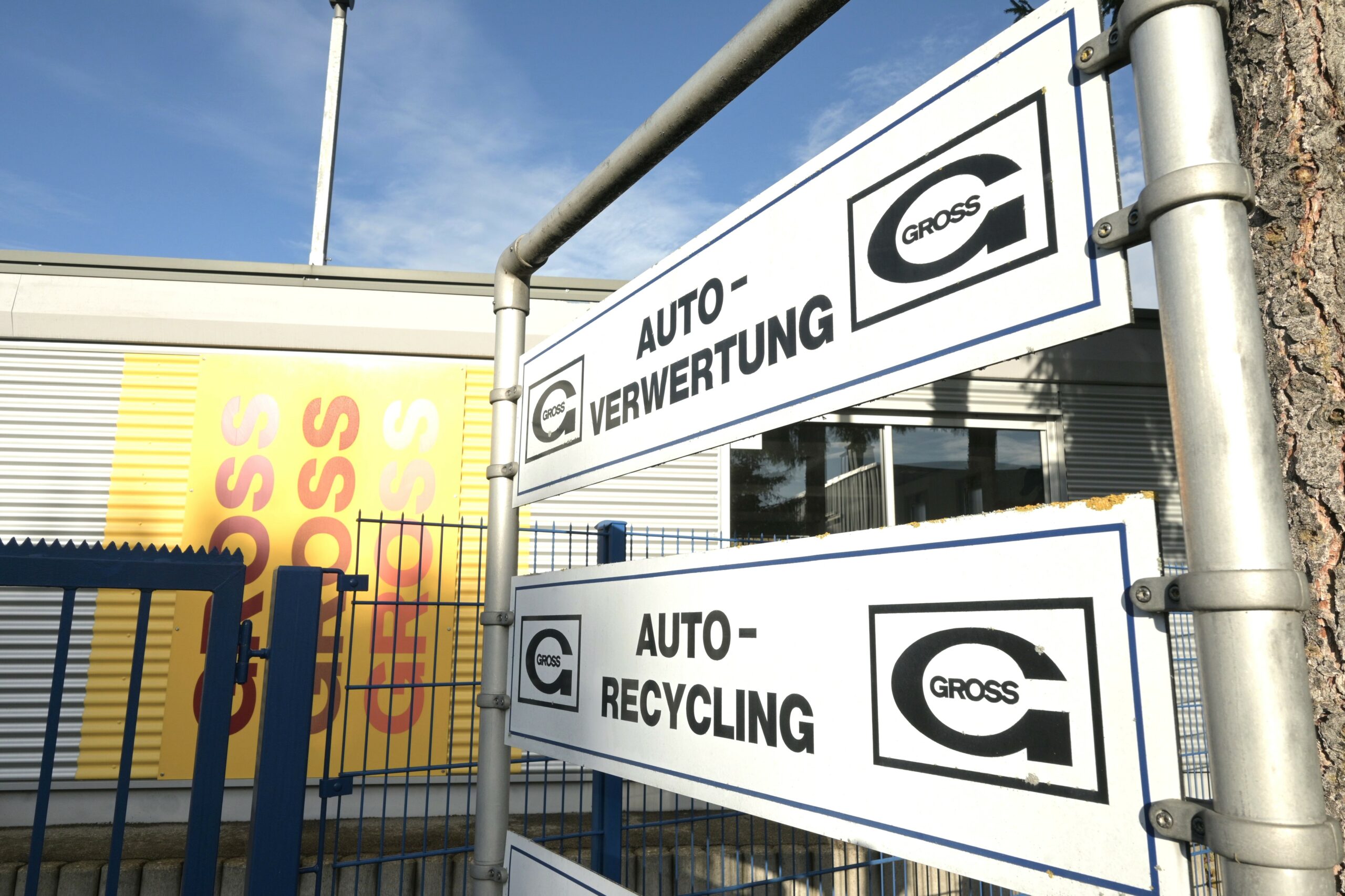 Schilder für Autoverwertung und Recycling neben einem Gebäude mit gelbem Schild und blauen Zaun bei Sonnenschein.
