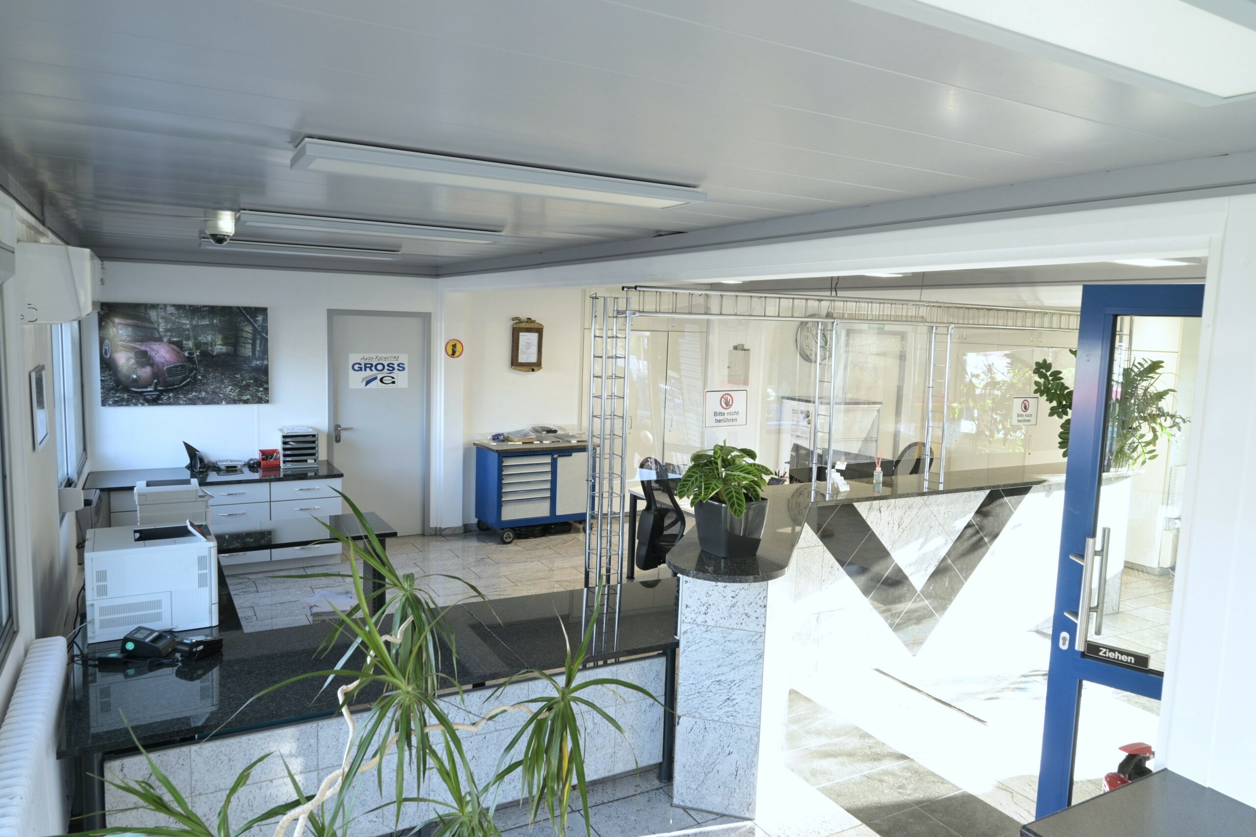 Helles Büro mit Empfangstresen, Pflanzen, Druckern und geschlossener Tür zur Werkstatt.