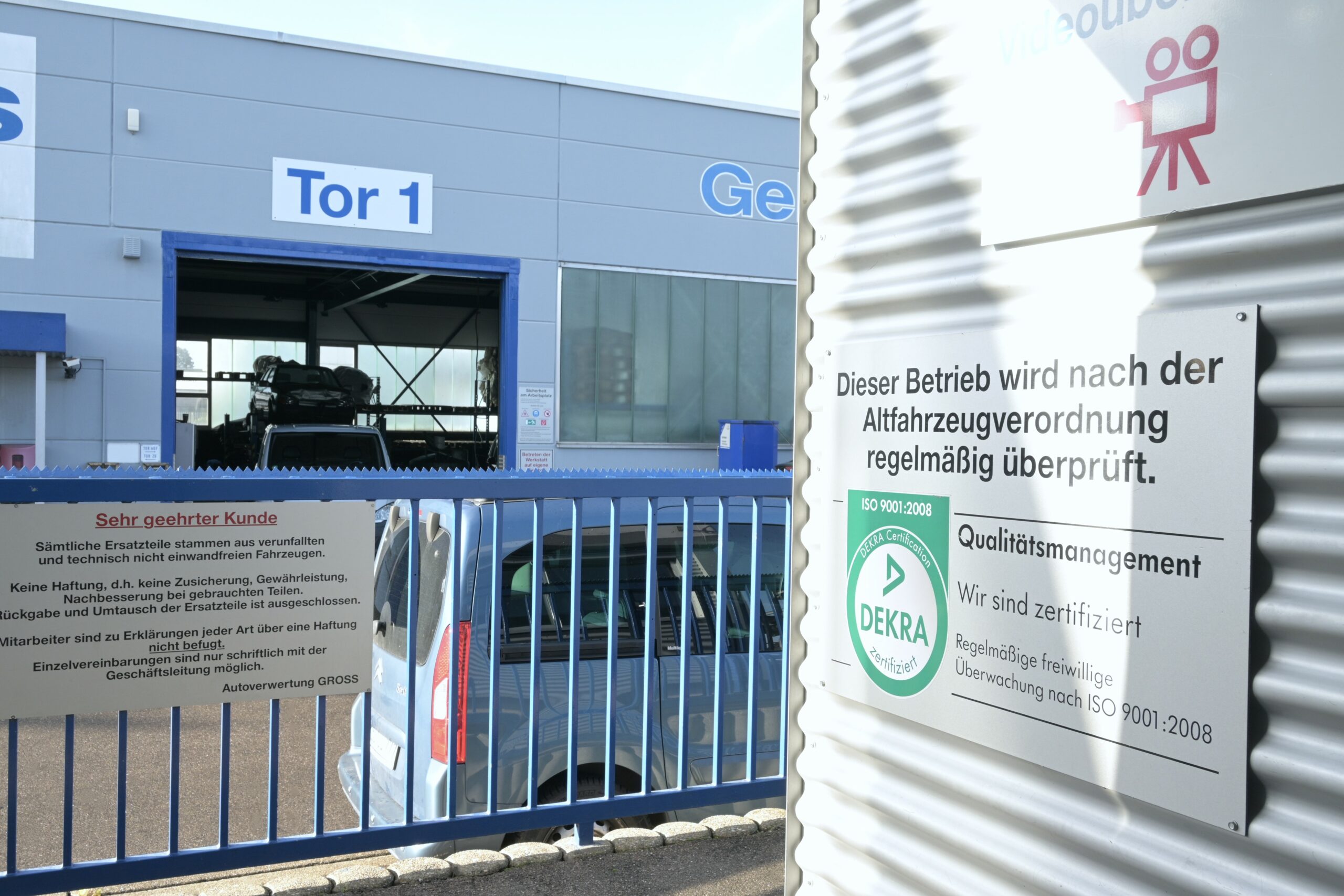 Autowerkstatt-Einfahrt mit Tor 1, DEKRA- und ISO-zertifizierte Prüfungshinweise an der Wand, blauer Zaun im Vordergrund.