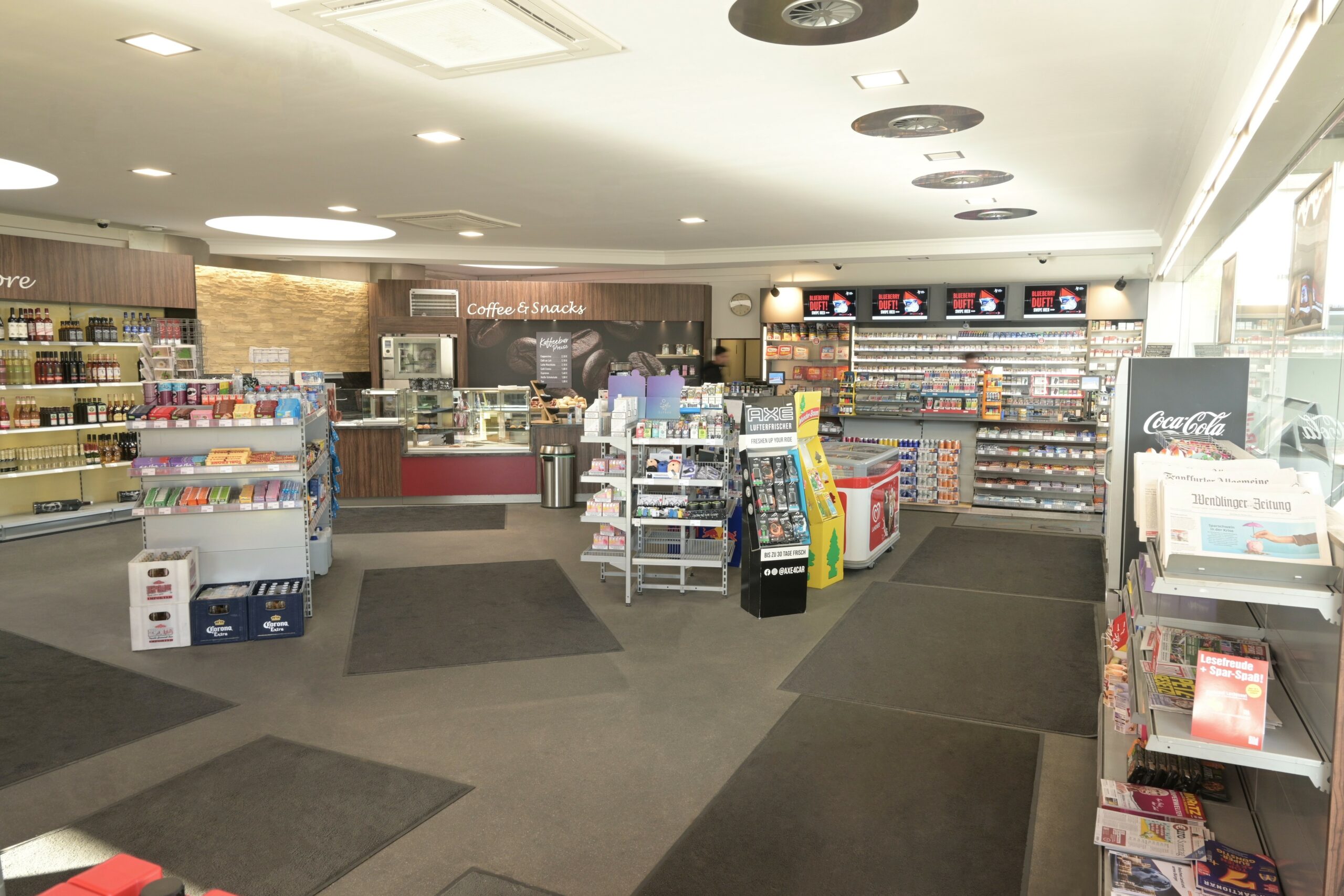 Ein heller Laden mit Regalen voller Snacks, Getränken und Zeitschriften. Ein Bereich für Kaffee und Snacks ist sichtbar.