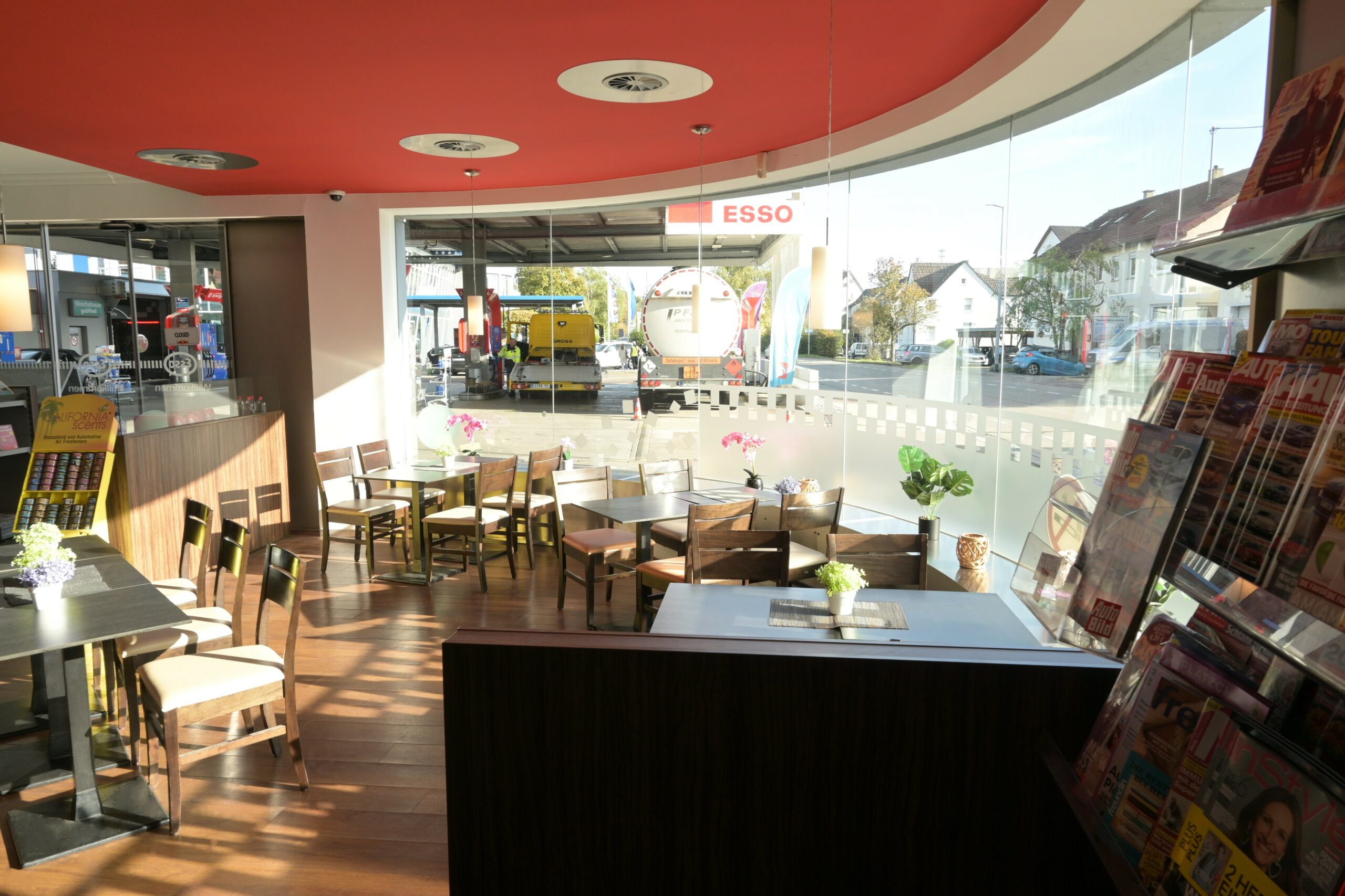 Sitzecke mit Holztischen und Stühlen in einer Tankstellenlounge, durch Fenster Blick auf Zapfsäulen und vorbeifahrendes Auto.