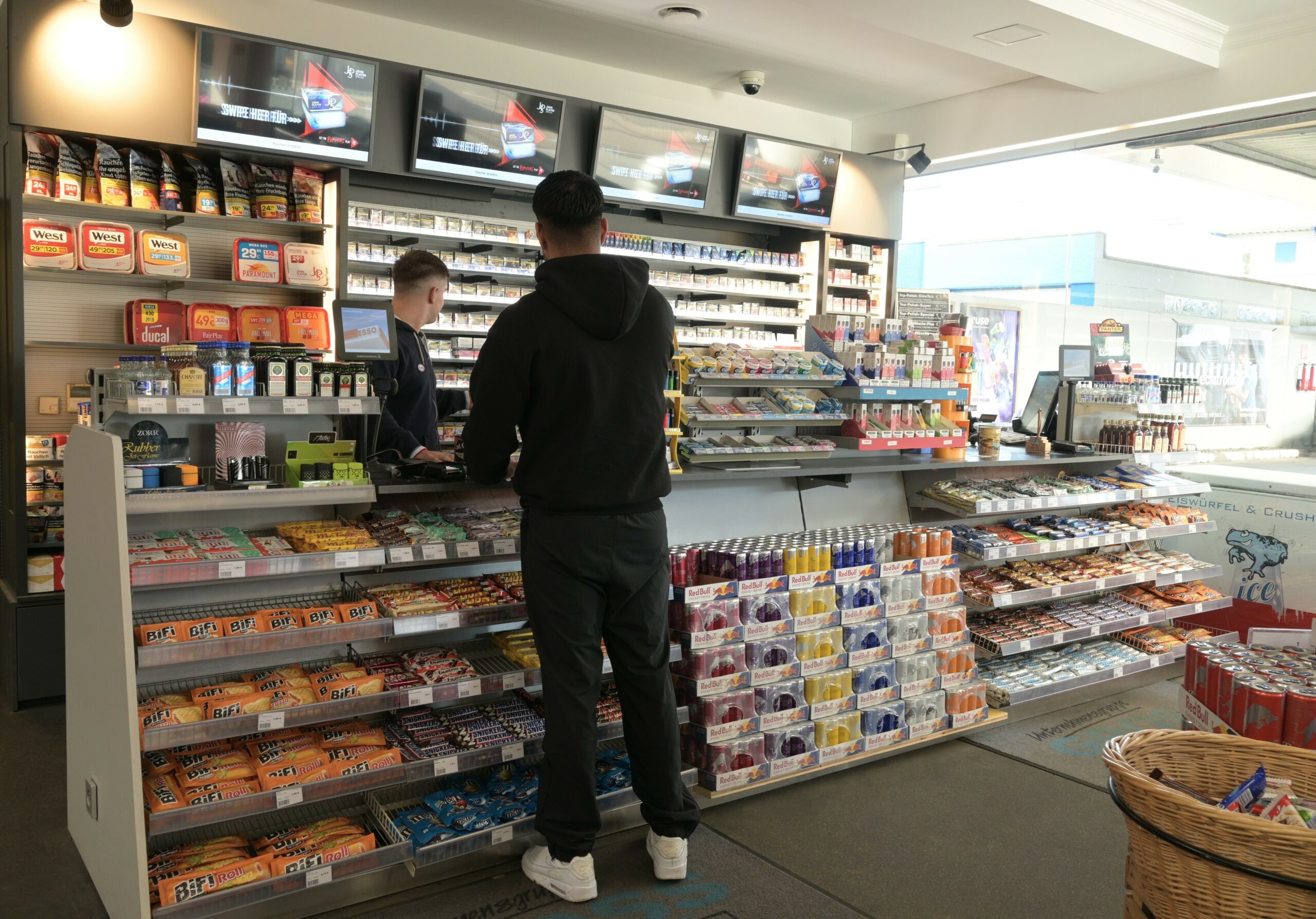 Ein Mann kauft in einem gut gefüllten Kiosk mit Snacks, Getränken und Tabakwaren. Verkäufer steht an der Kasse.