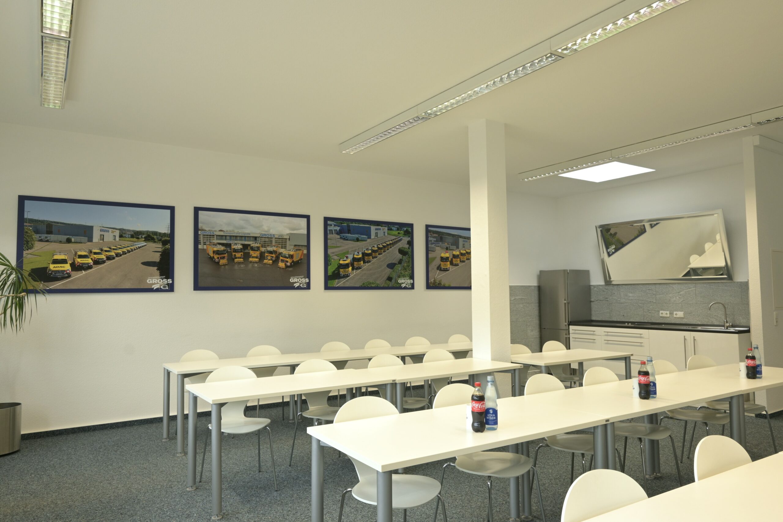 Moderner Schulungsraum mit Tischen, Stühlen und drei Wandbildern von gelben Fahrzeugen. Kühlschrank und Spiegel im Hintergrund.