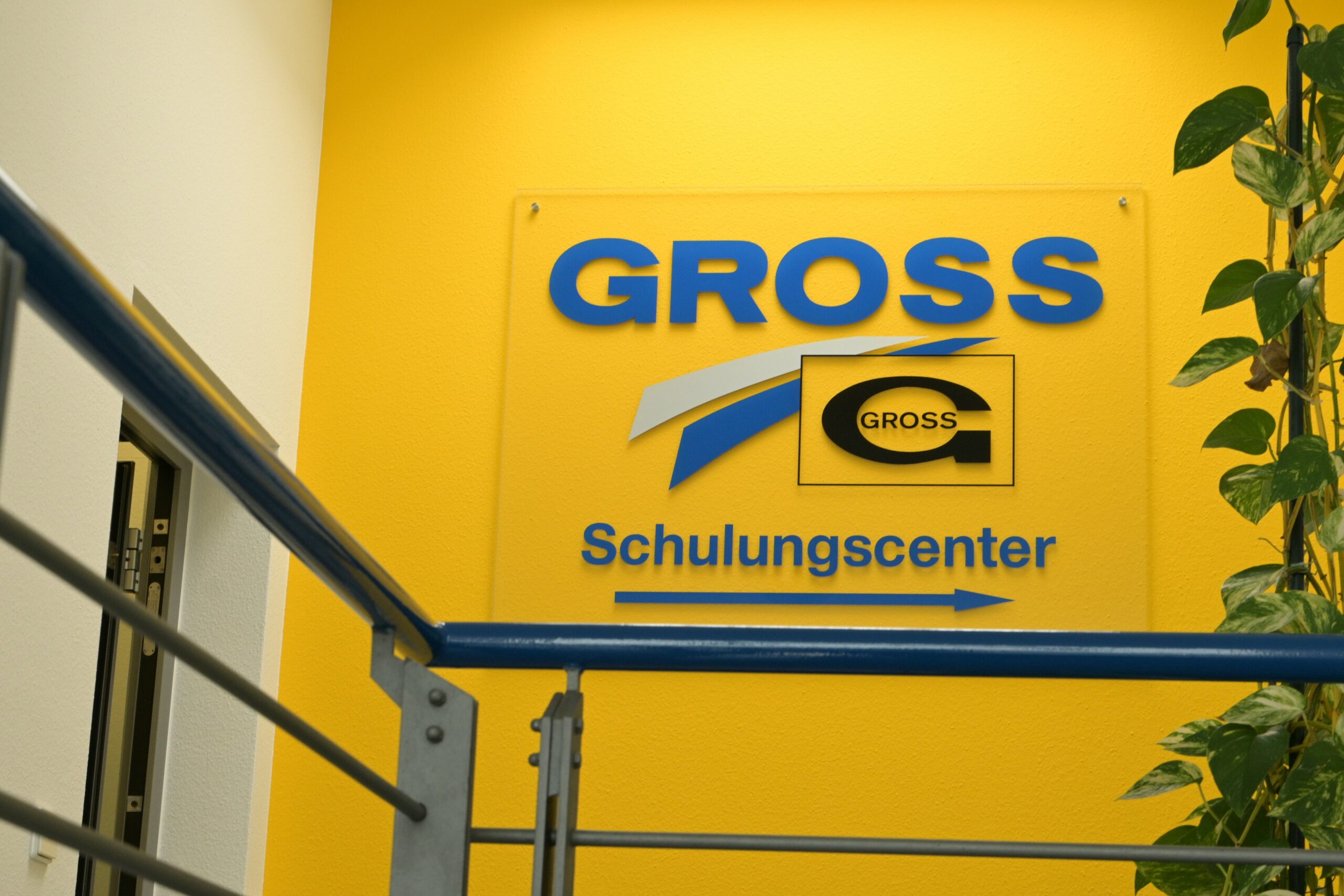 Gelbes Schild mit blauem Schriftzug „Gross Schulungscenter“ neben einer Pflanze und einem Geländer in einem Gebäude.