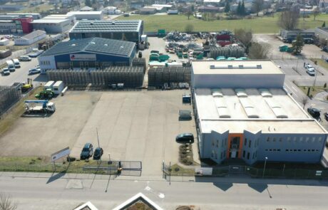 Luftbild eines Industriekomplexes mit zwei großen Hallen, geparkten Fahrzeugen und umliegender Landschaft bei sonnigem Wetter.