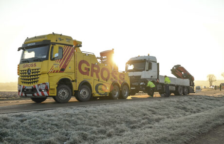 Gelber Abschleppwagen mit GROSS zieht weißen Lastwagen auf frostiger Straße bei Sonnenaufgang; zwei Personen arbeiten daran.
