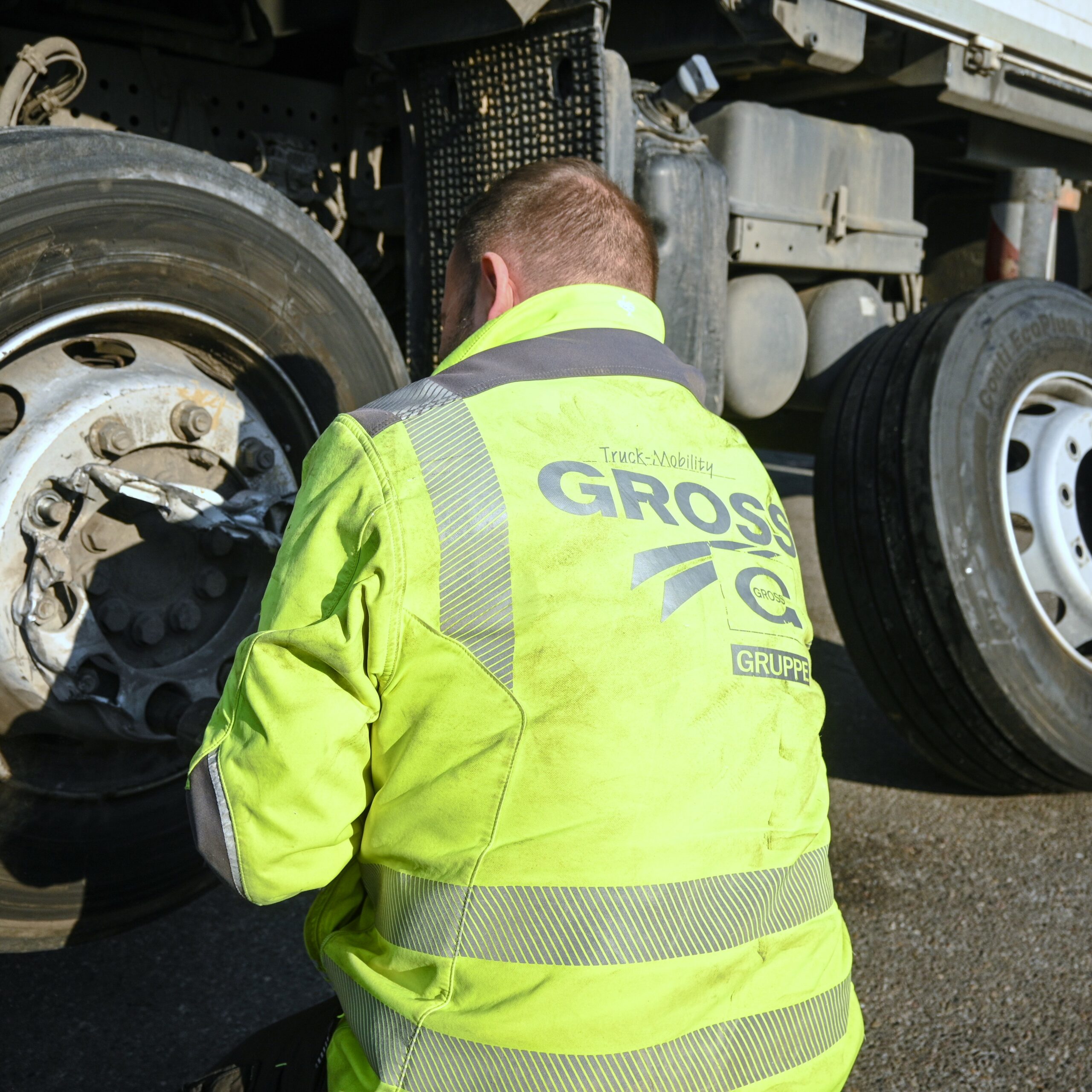 Mann in gelber Warnweste repariert Lkw-Rad auf einer Straße, Nahaufnahme vom Rücken.
