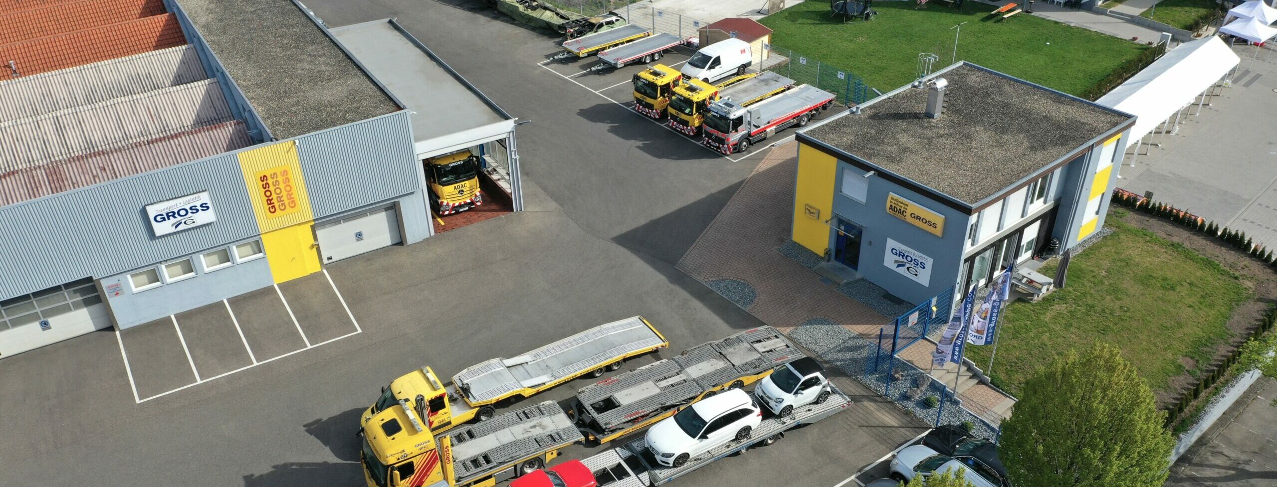 Luftbild einer Autofirma mit gelben Abschleppwagen und zwei Gebäuden im Hof. Mehrere Autos auf Transporter geladen.