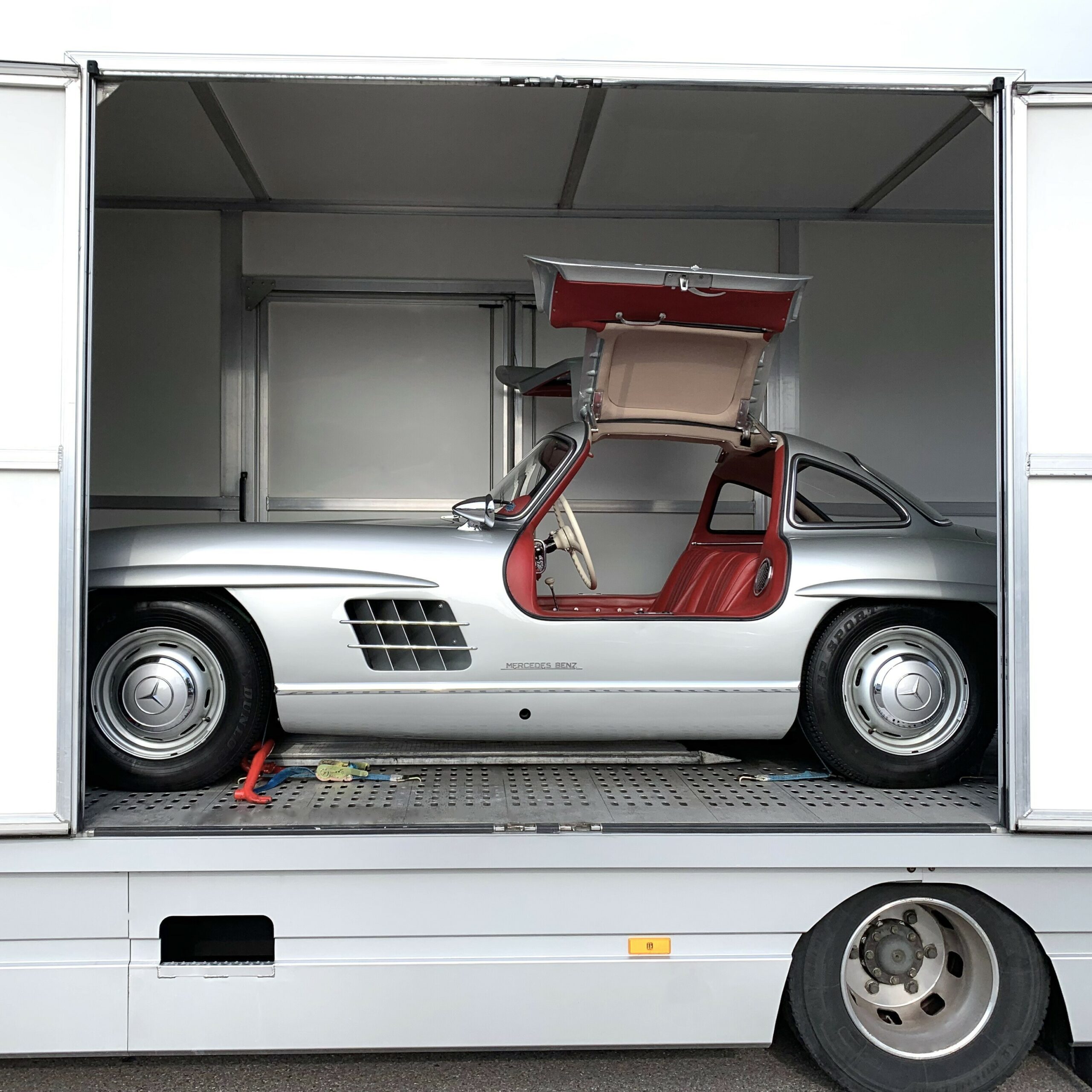 Silberner Oldtimer mit Flügeltür in einem Transporter, Tür geöffnet, rote Innenausstattung sichtbar.