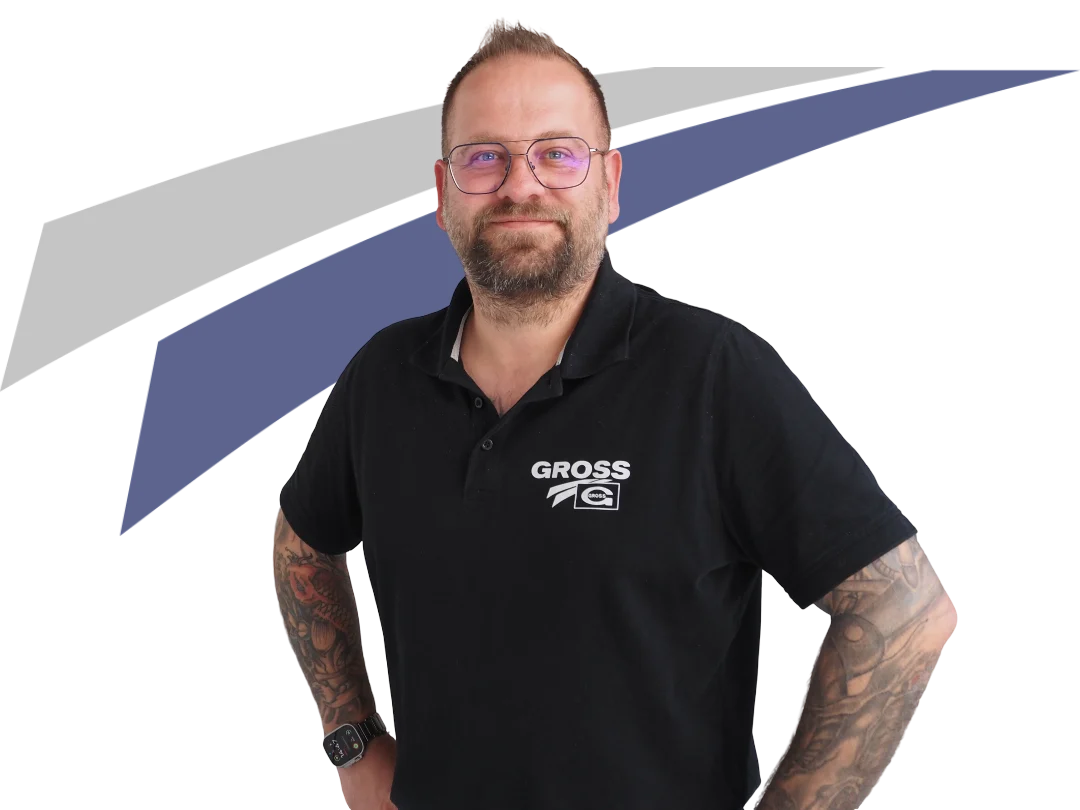 Mann mit Bart und Tätowierungen in schwarzem Polo-Shirt mit Aufdruck „GROSS“, lächelt vor neutralem Hintergrund.