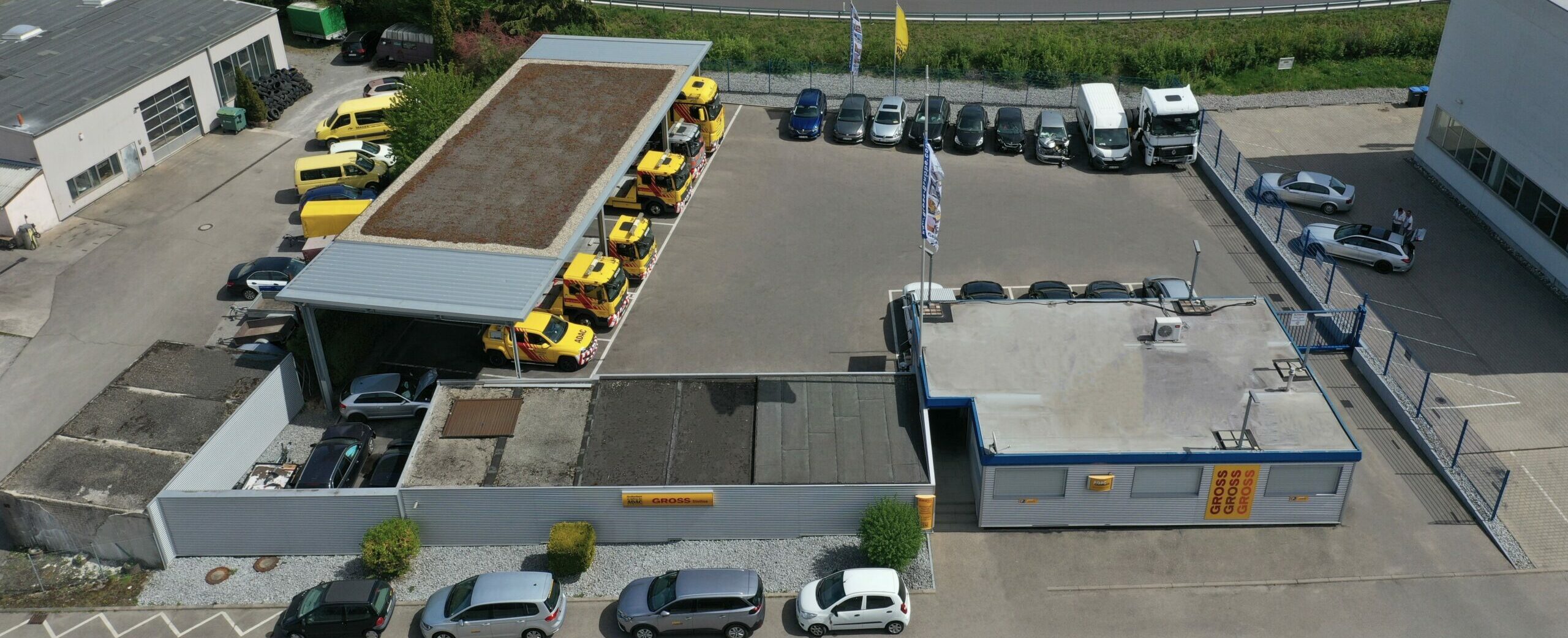 Luftaufnahme eines Parkplatzes mit gelben Einsatzfahrzeugen unter Dach, umgeben von Gebäuden und parkenden Autos.