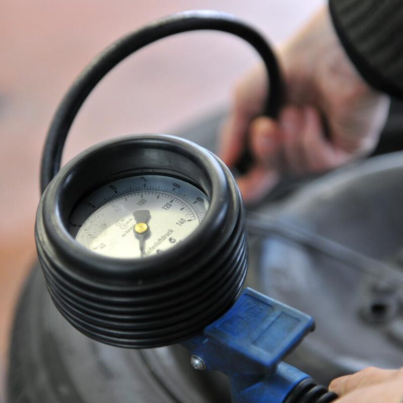 Nahaufnahme eines Reifendruckmessgeräts, das auf einen Autoreifen gerichtet ist. Eine Hand hält den Messschlauch.