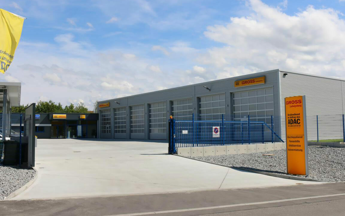 Kfz-Werkstatt mit blauem Zaun und gelbem Schild bei sonnigem Wetter, mehrere Garagentore, klare Zufahrt.